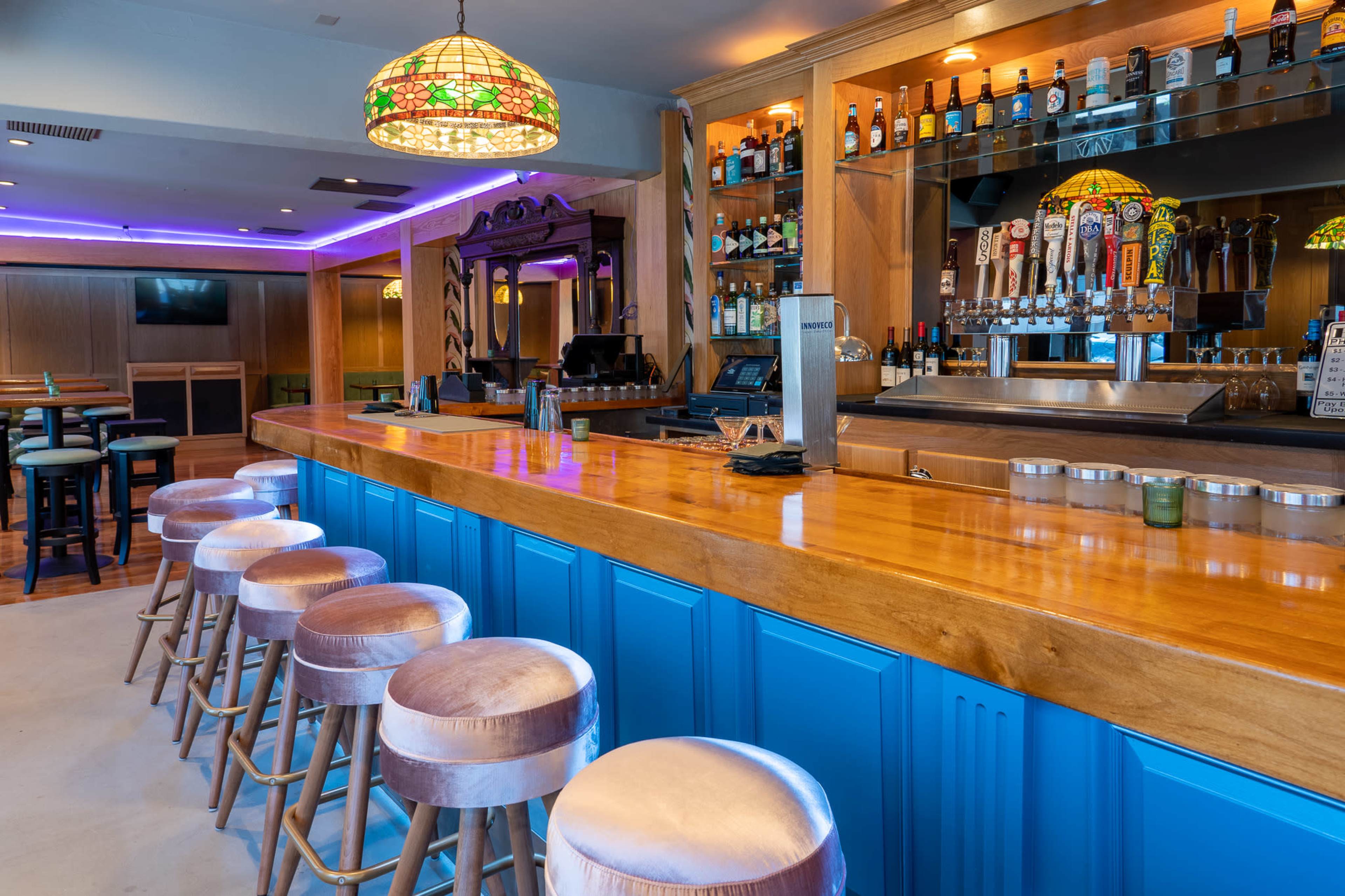 A brightly lit bar features a polished wooden counter with several padded stools and shelves lined with various bottles and taps.