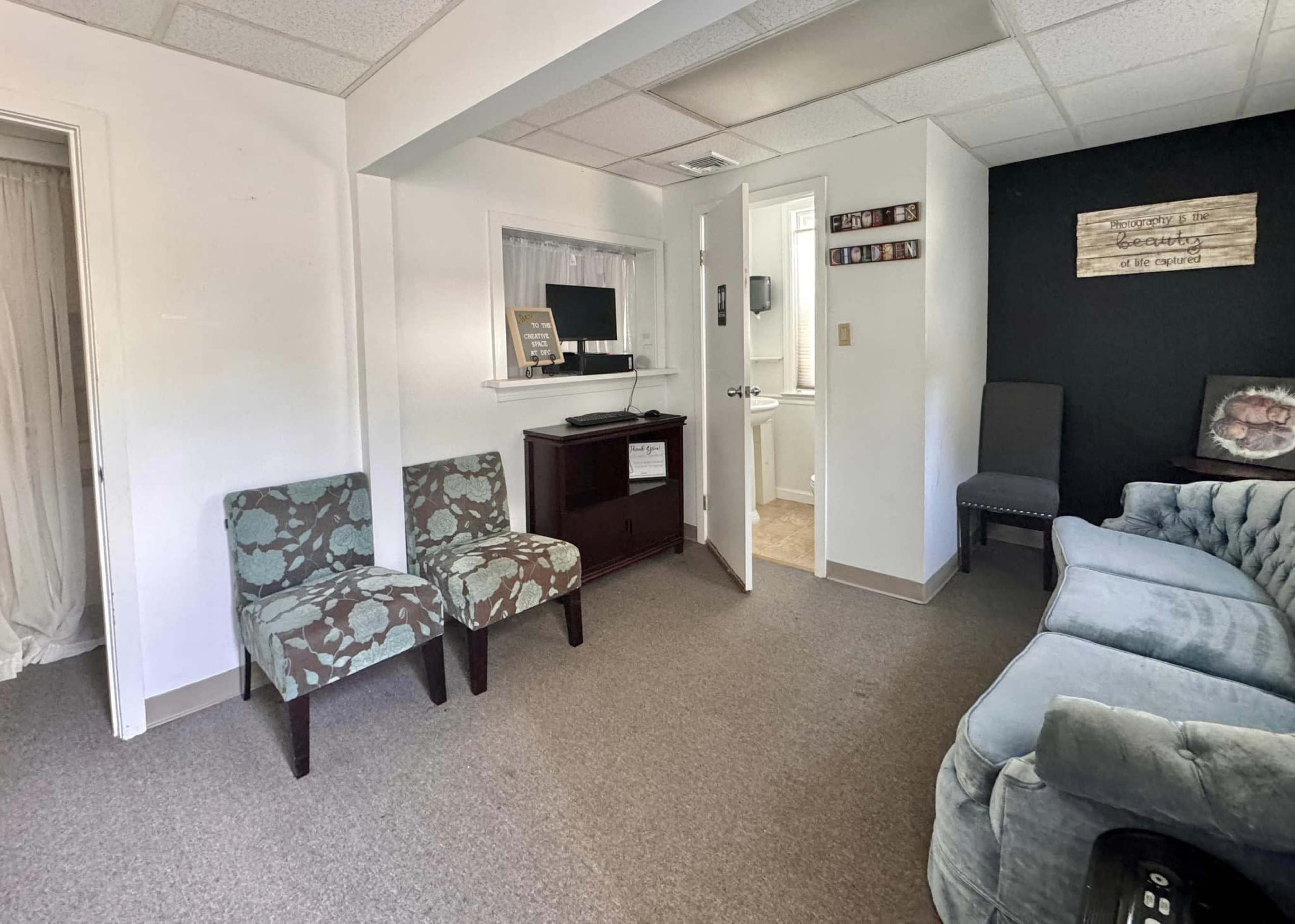 The image shows a small living area with two patterned chairs, a wooden cabinet, and a gray sofa, with a doorway leading to a bathroom and a wall displaying decorations.