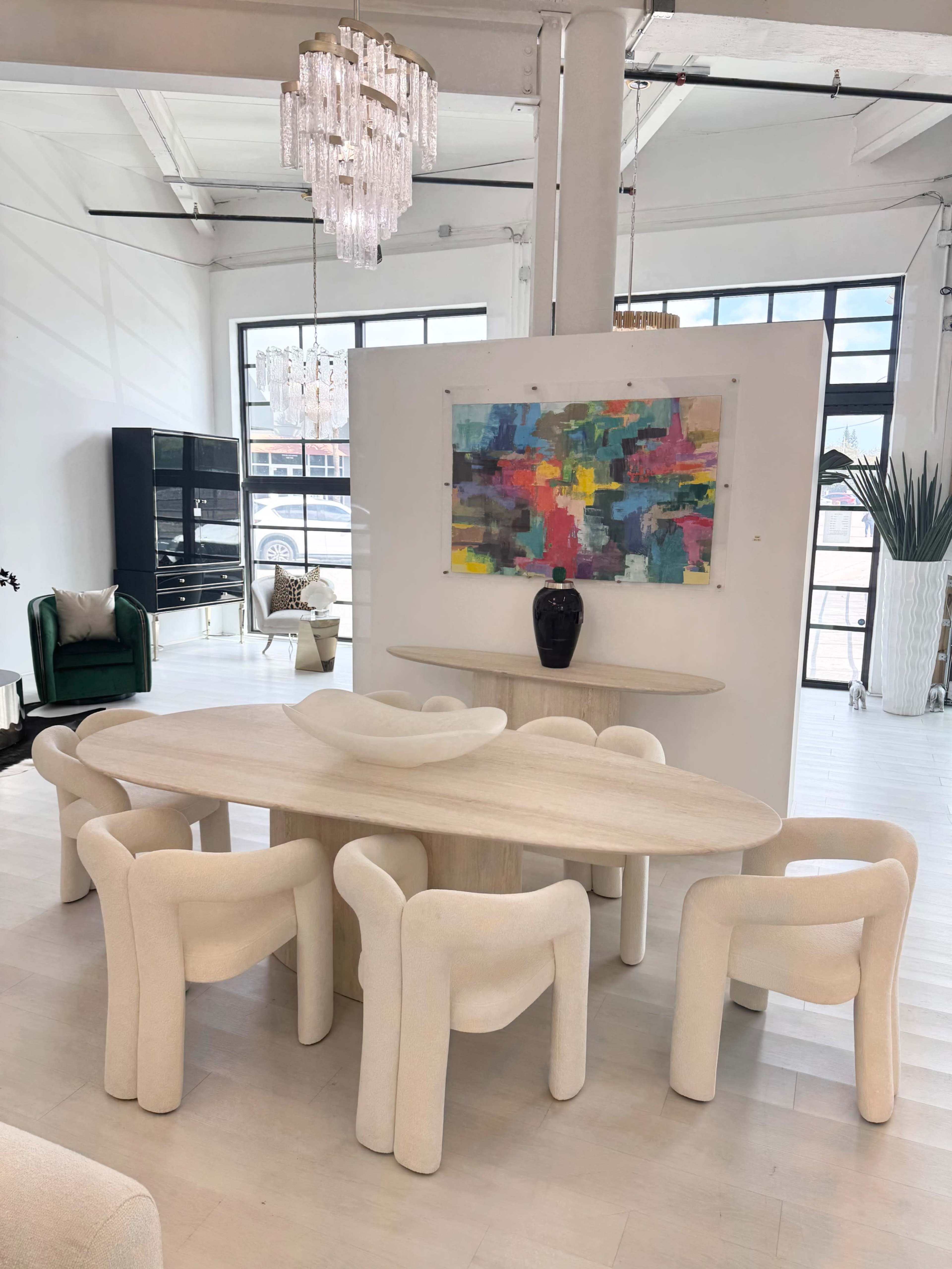 A modern dining area features a light-colored oval table surrounded by plush, rounded chairs, with a colorful abstract painting displayed on the wall.