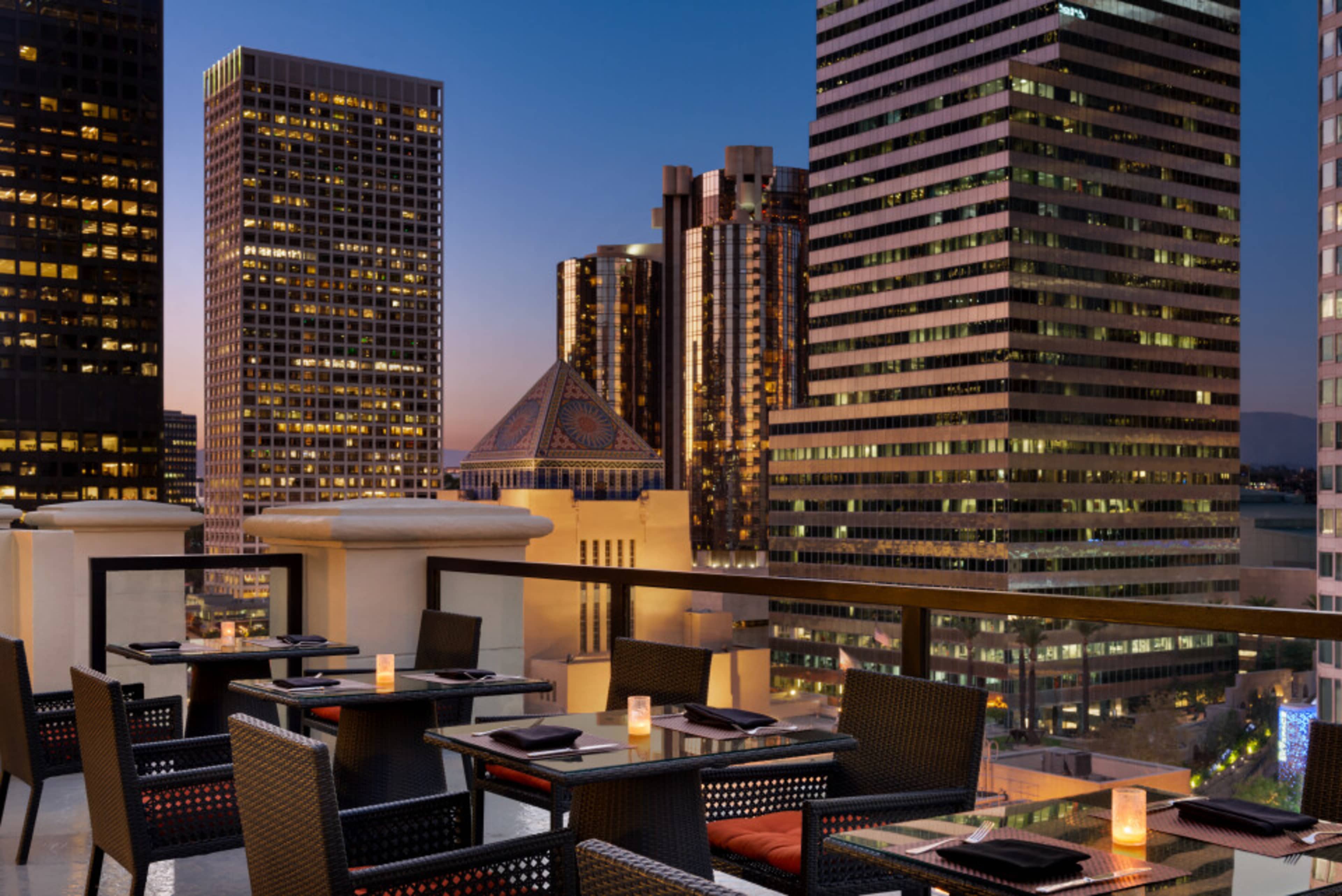 A rooftop dining area features tables with candles, overlooking a city skyline at dusk.