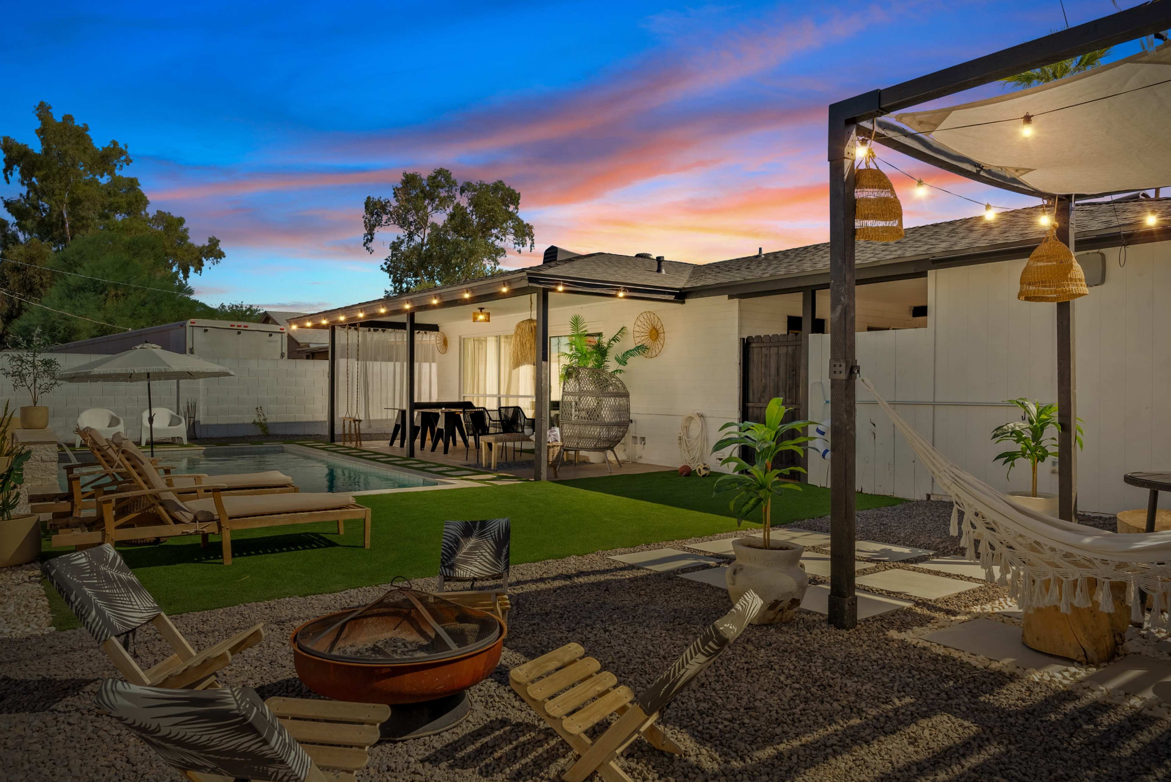The image features a well-lit backyard at dusk, with a swimming pool, lounge chairs, a dining area, and decorative plants set against a colorful sunset sky.
