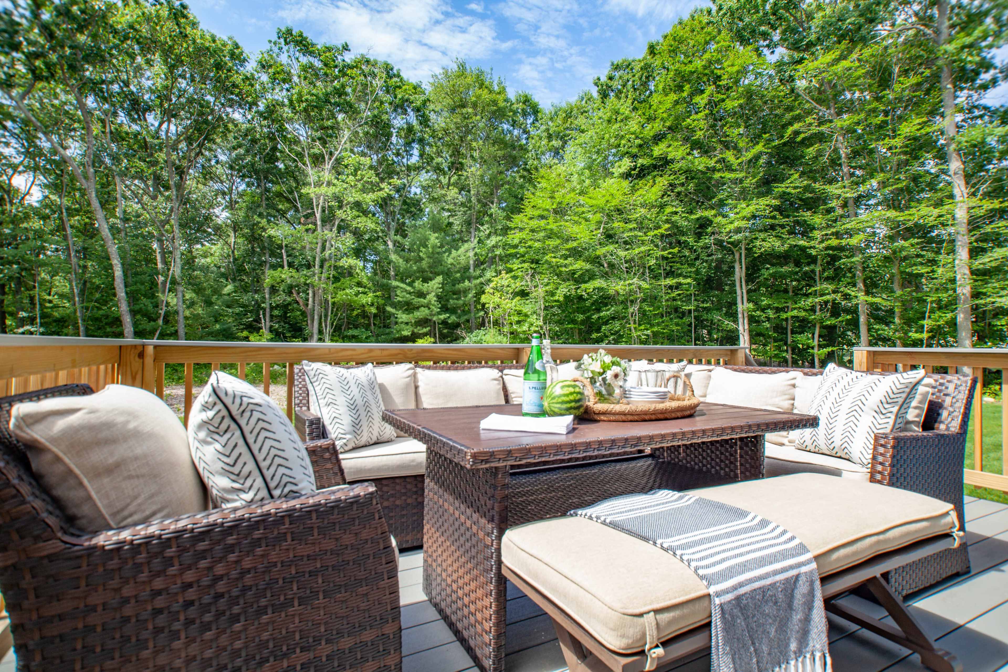 A furnished outdoor deck features a woven seating arrangement with cushions, a central coffee table, and potted plants surrounded by trees.