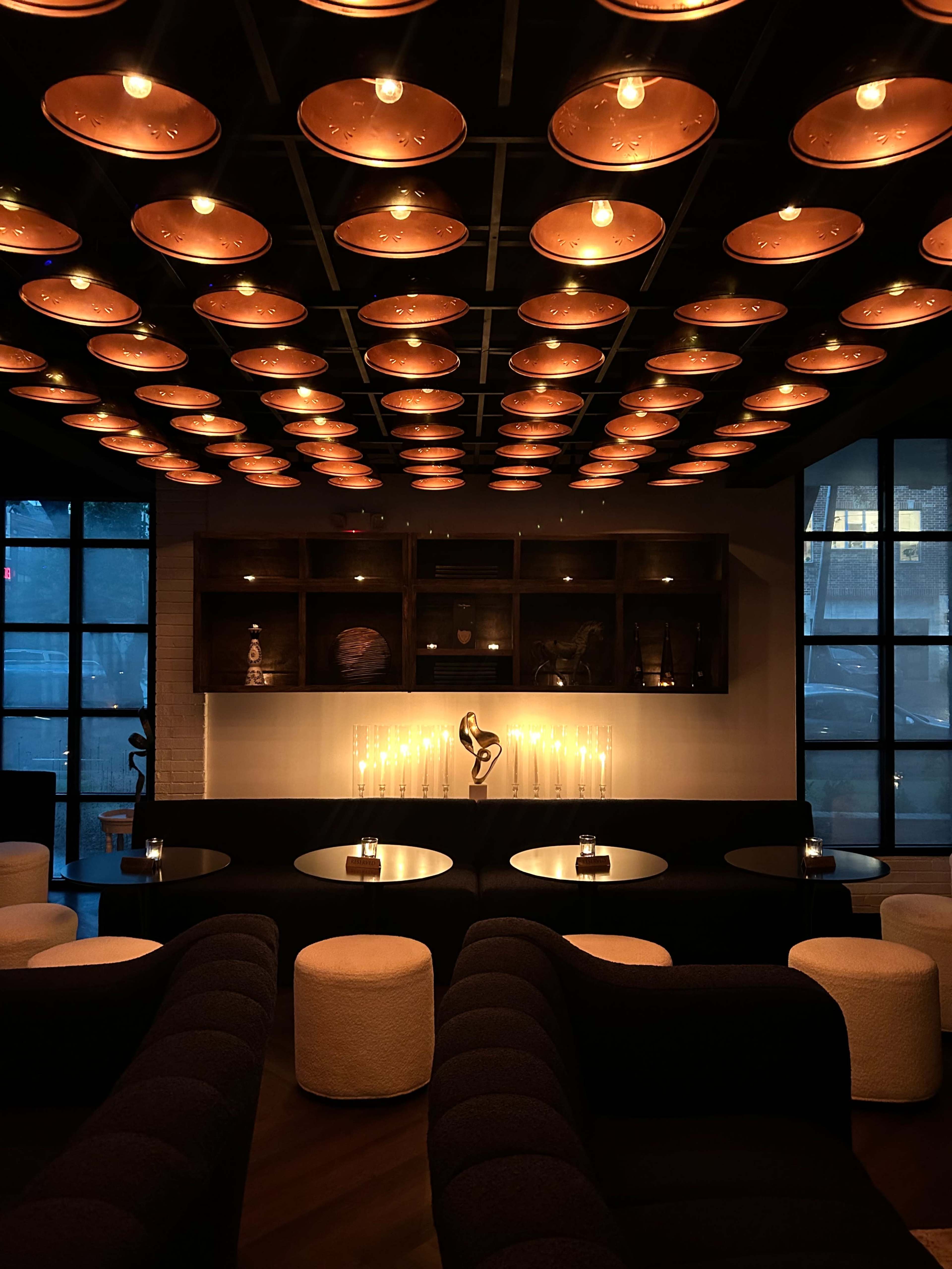 The image shows a modern lounge with a dark ceiling featuring round light fixtures and a wall displaying shelves with decorative items, complemented by low tables and soft seating.
