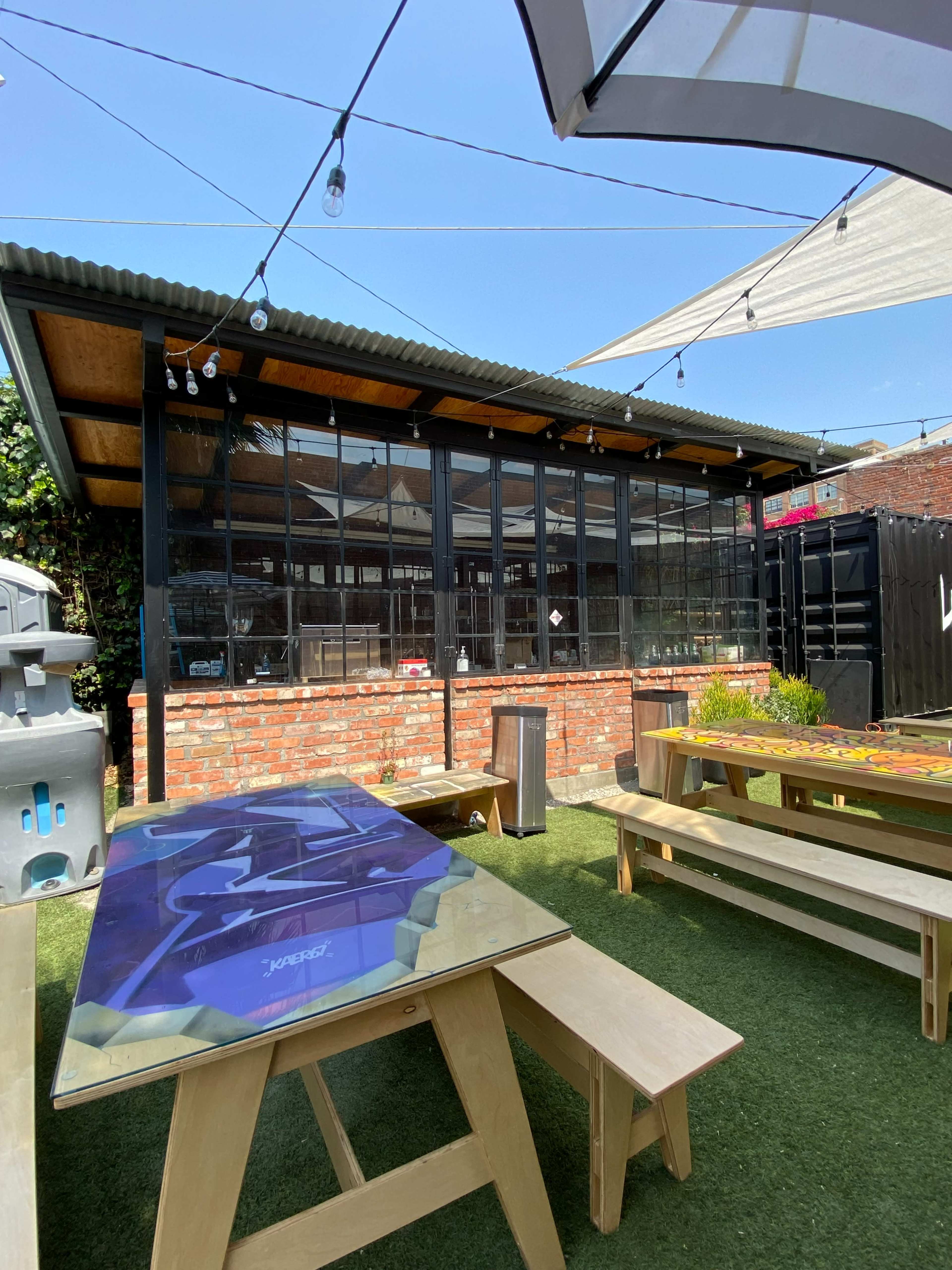 An outdoor dining area with wooden benches and tables set on grass, adjacent to a glass-walled building.
