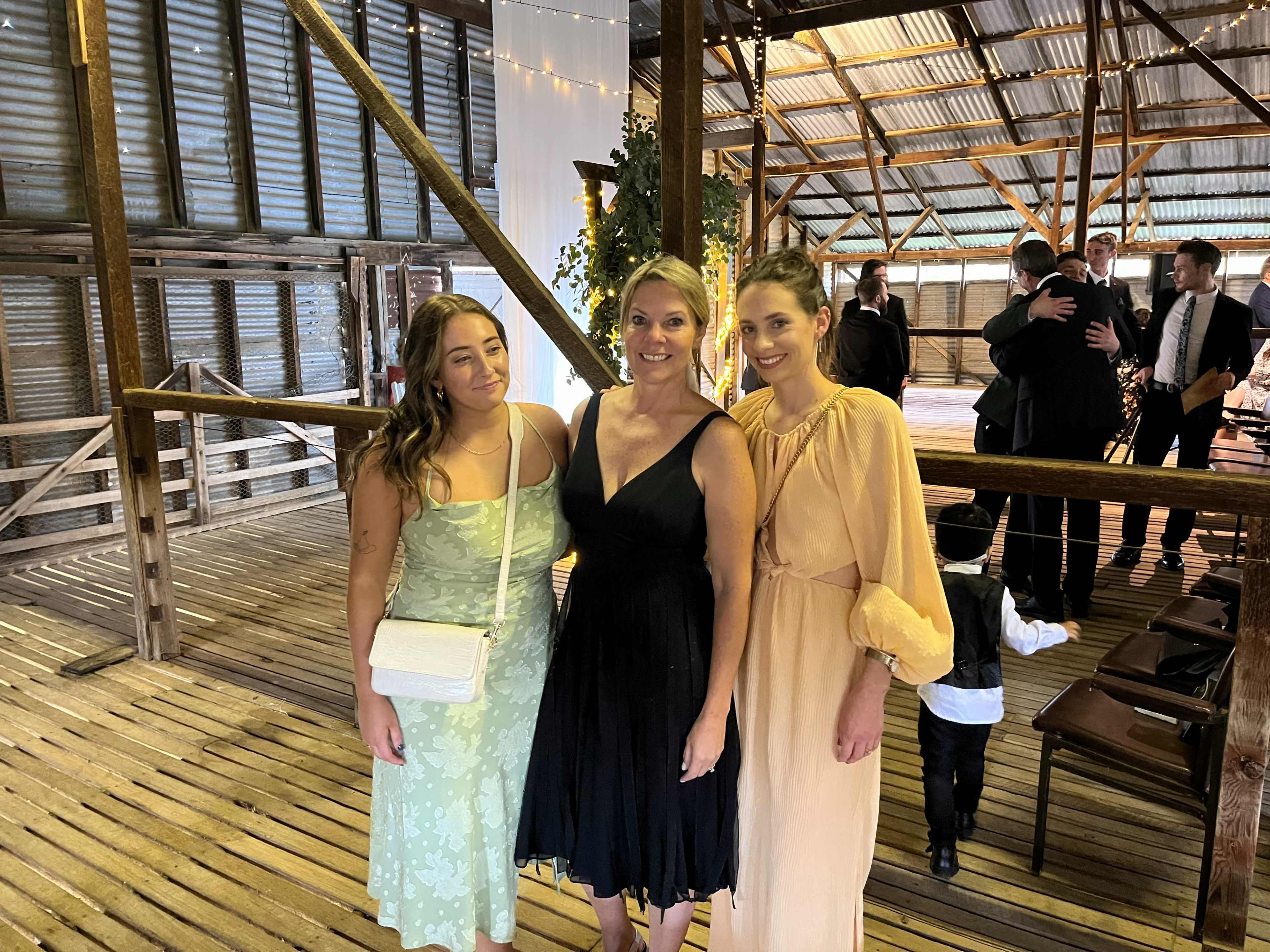 Three women pose together in a rustic barn setting at a social event.