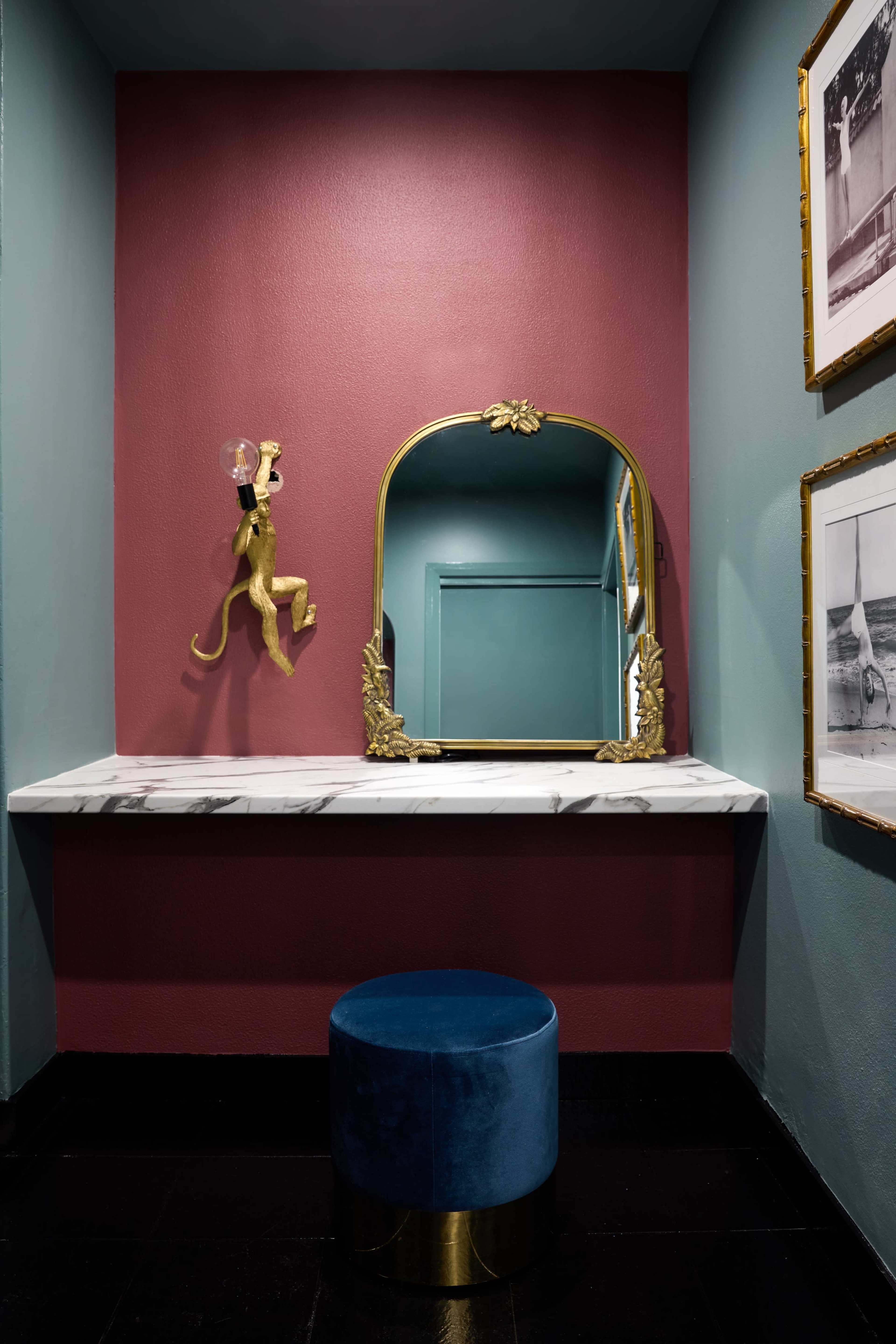 The image features a small bathroom with a maroon wall, a marble-topped vanity, an ornate gold-framed mirror, and a blue velvet stool.