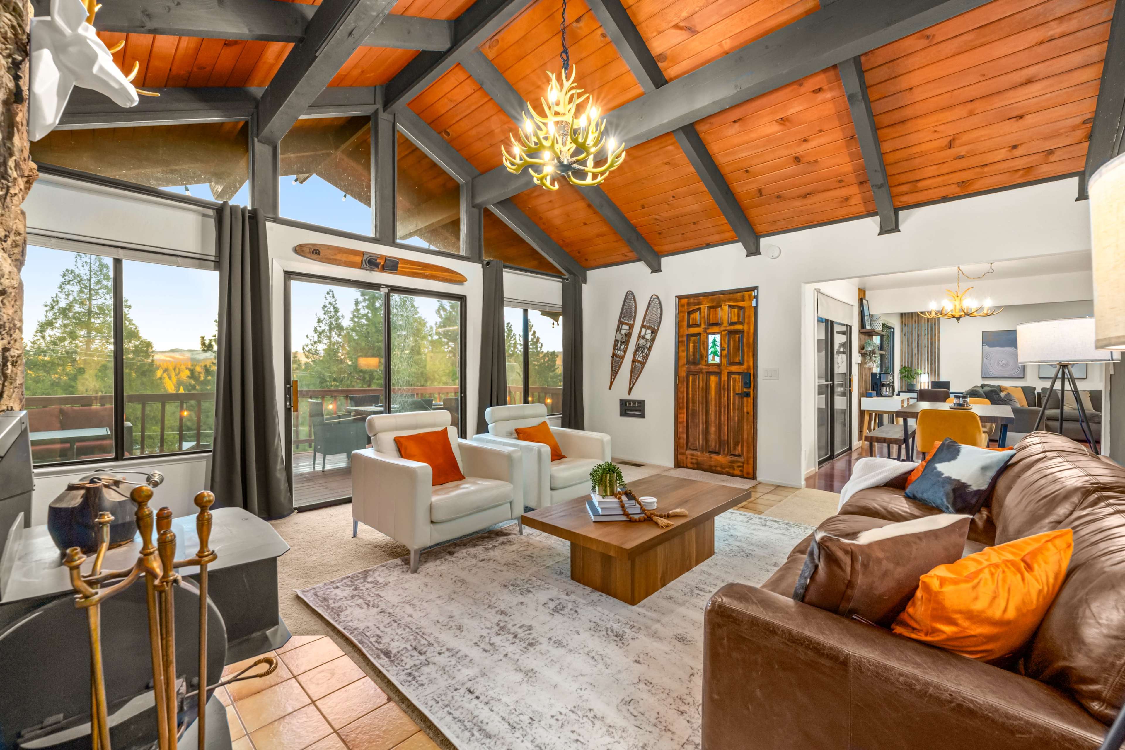 The living room features a high-beamed ceiling, large windows with a view of the outdoors, a central wooden coffee table, and a mix of seating arrangements.
