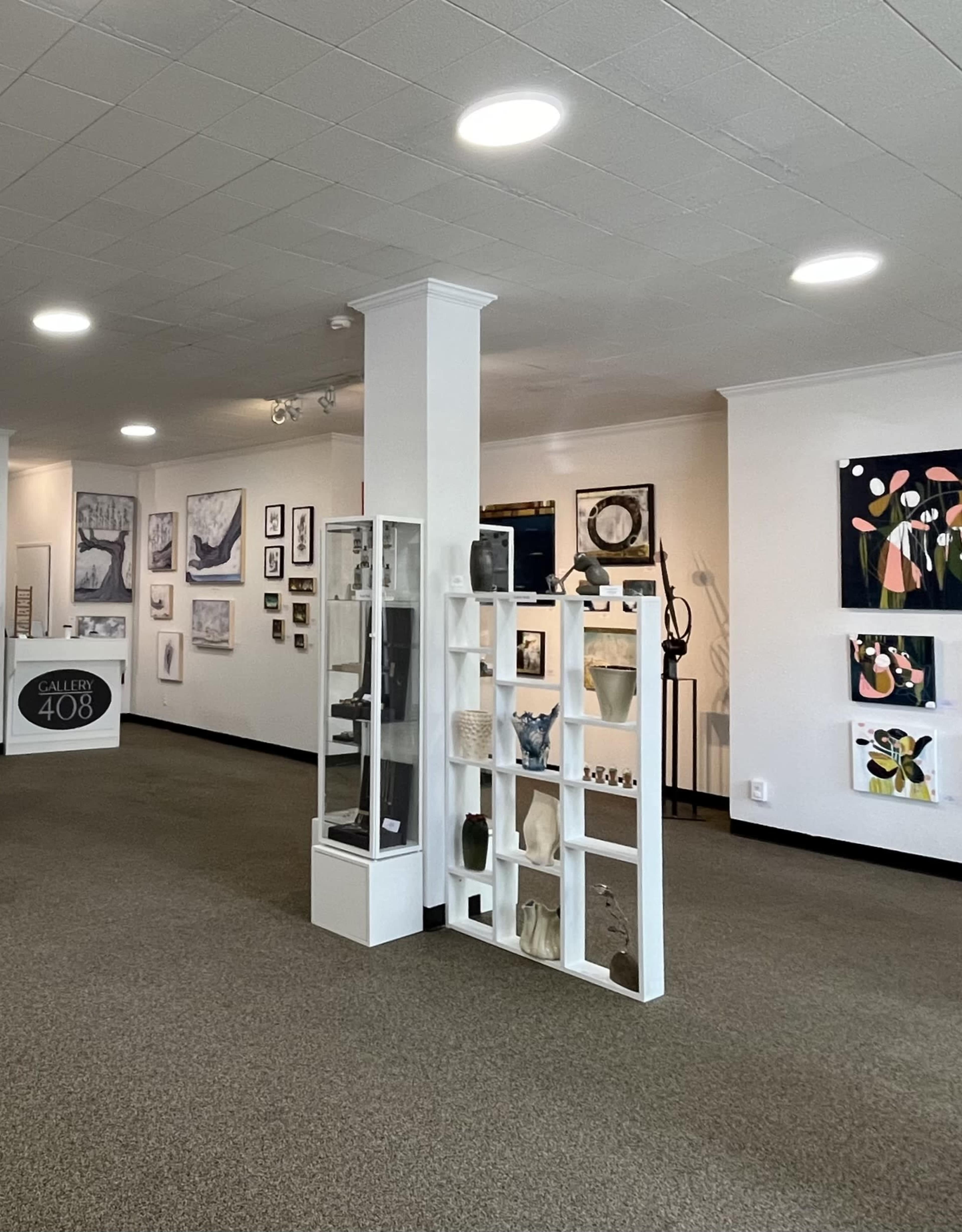 The image shows an art gallery interior with various framed artworks on the walls and a white display shelf featuring sculptures and ceramics.