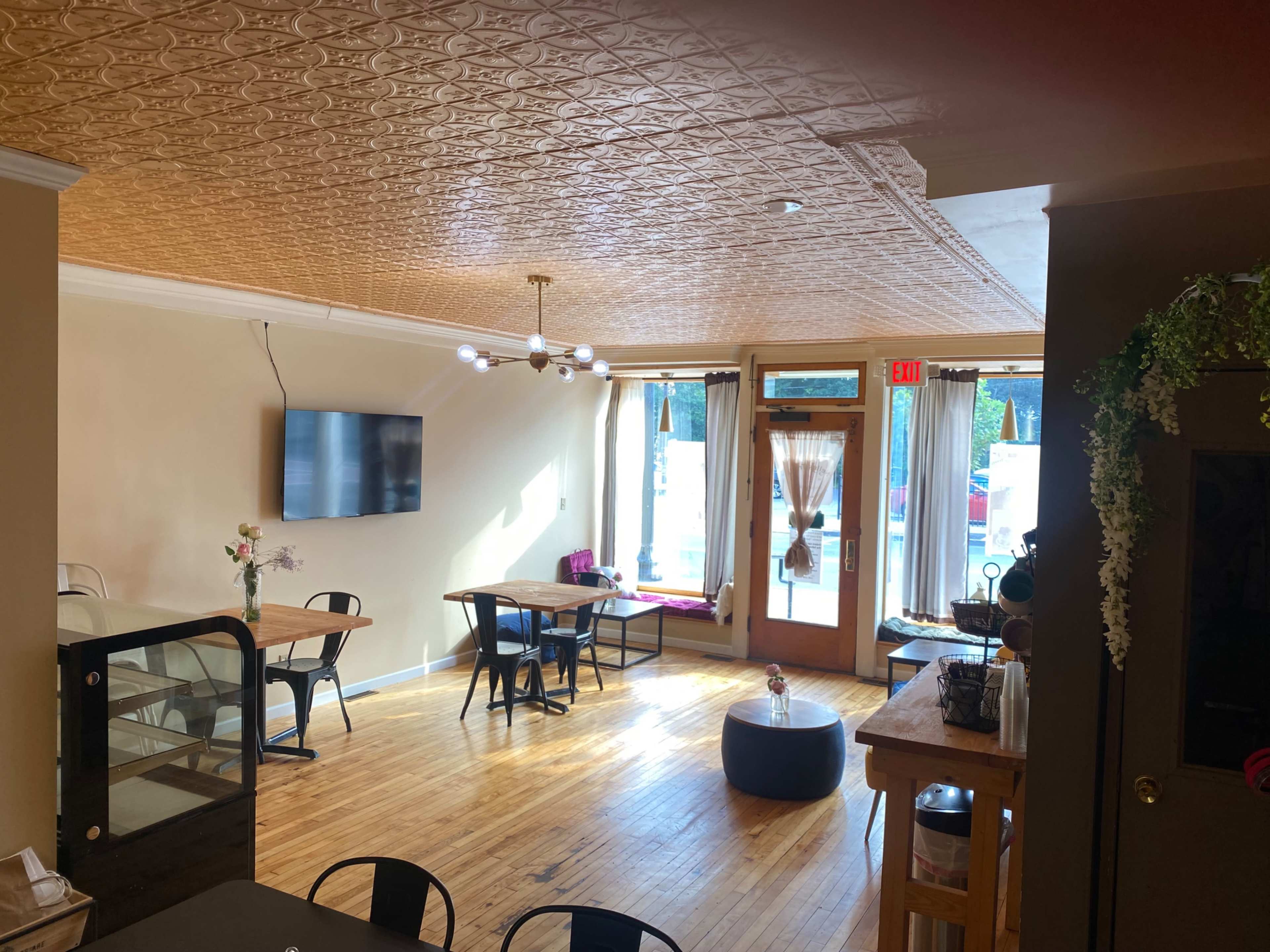 A brightly lit cafe interior with wooden floors, several tables, a television mounted on the wall, and large windows letting in natural light.