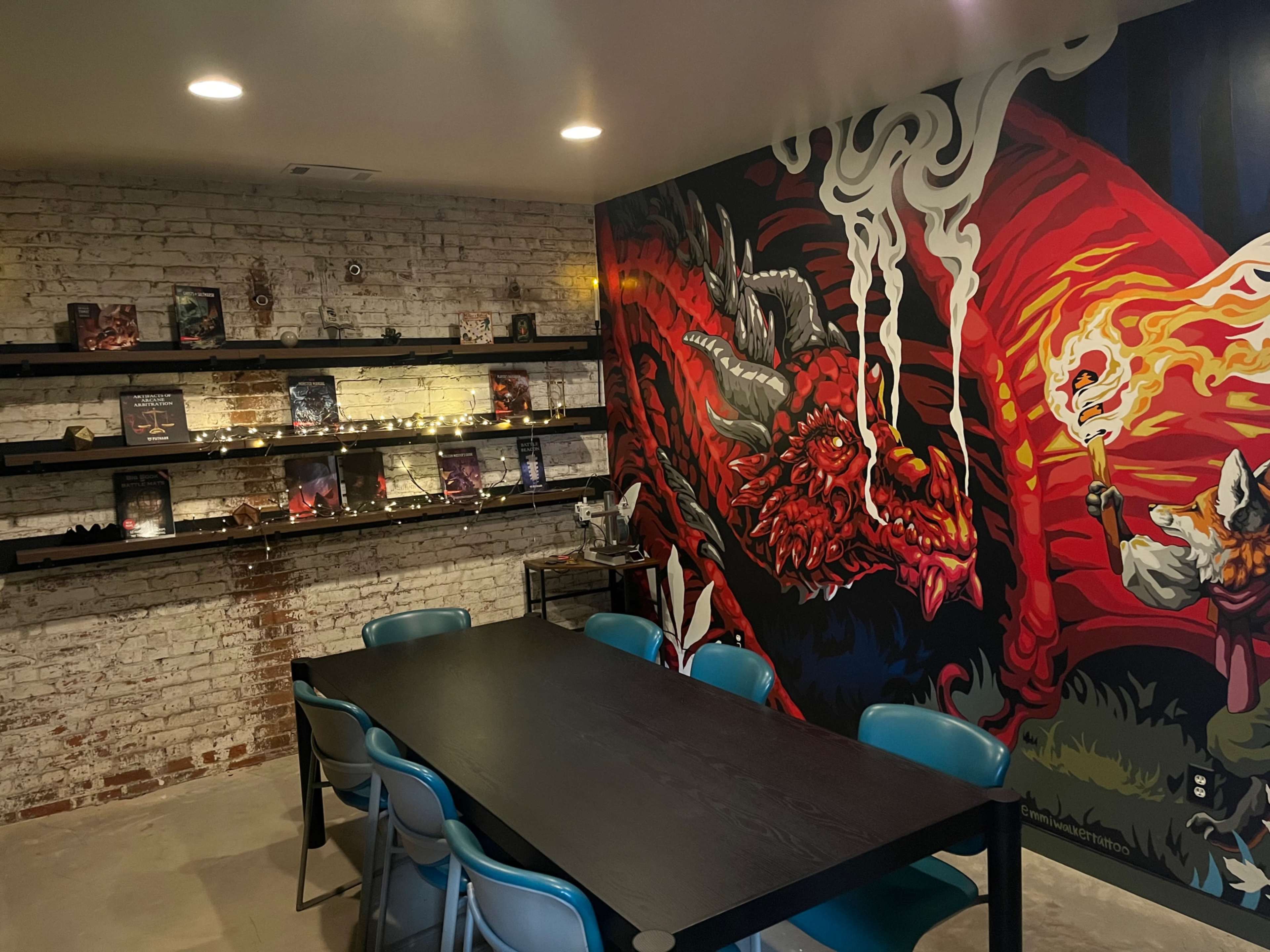 The image shows a room with a large, colorful mural of a dragon on one wall, shelves displaying various books and collectibles, and a black table surrounded by teal chairs.