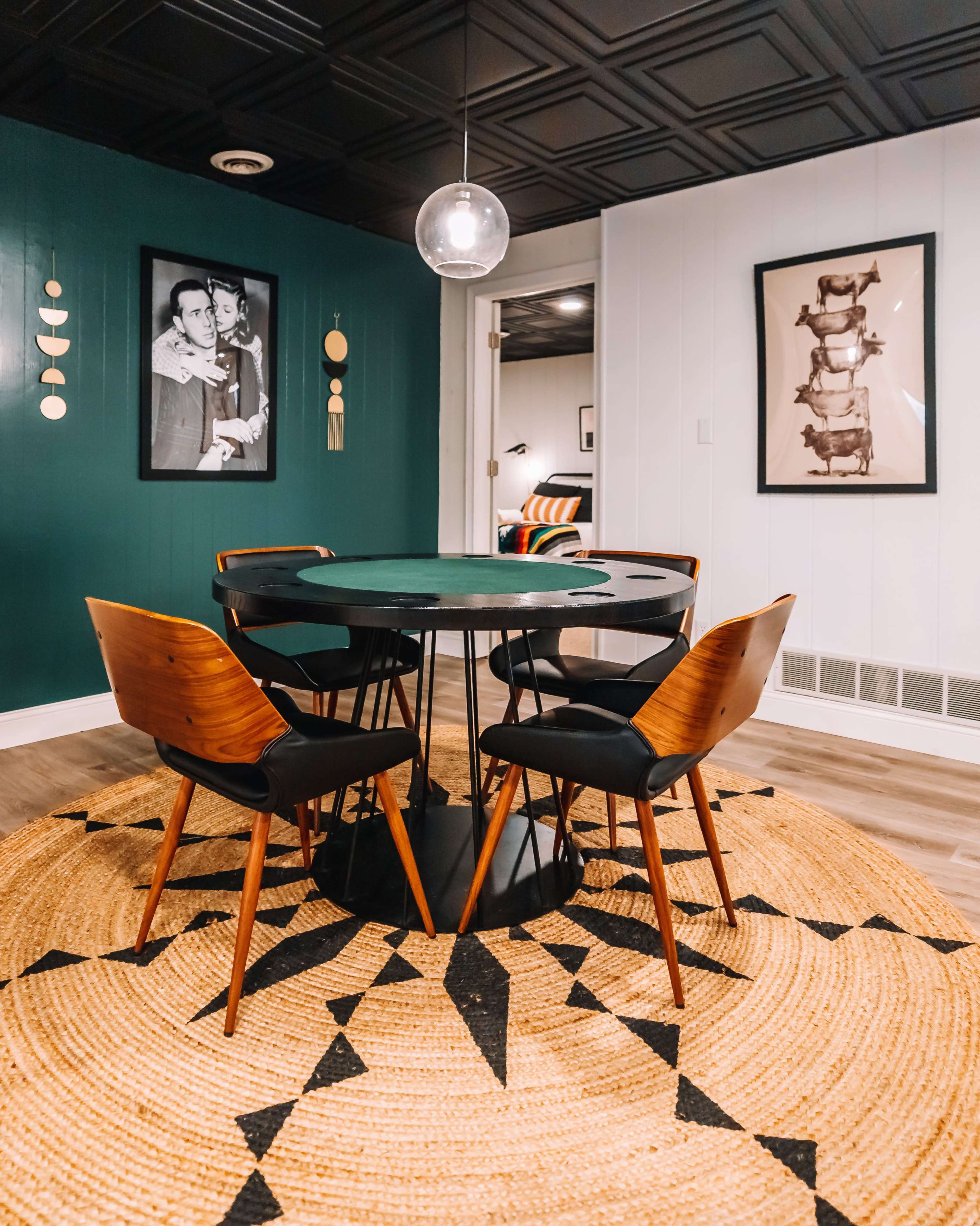 A round dining table with four wooden chairs is positioned on a patterned rug in a room with dark green walls and framed artwork.