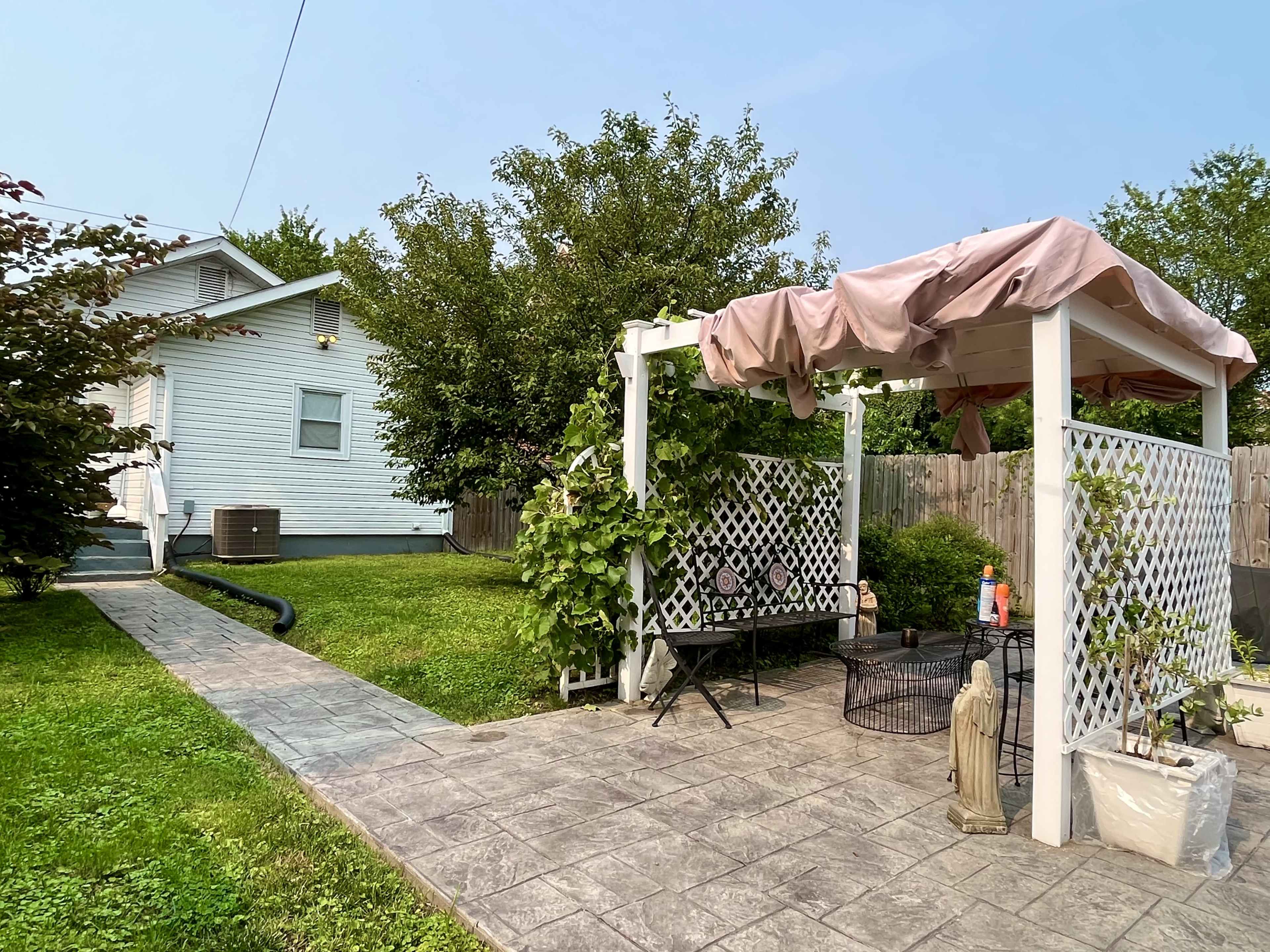 A patio with a covered seating area and surrounding greenery is located beside a house in a residential yard.