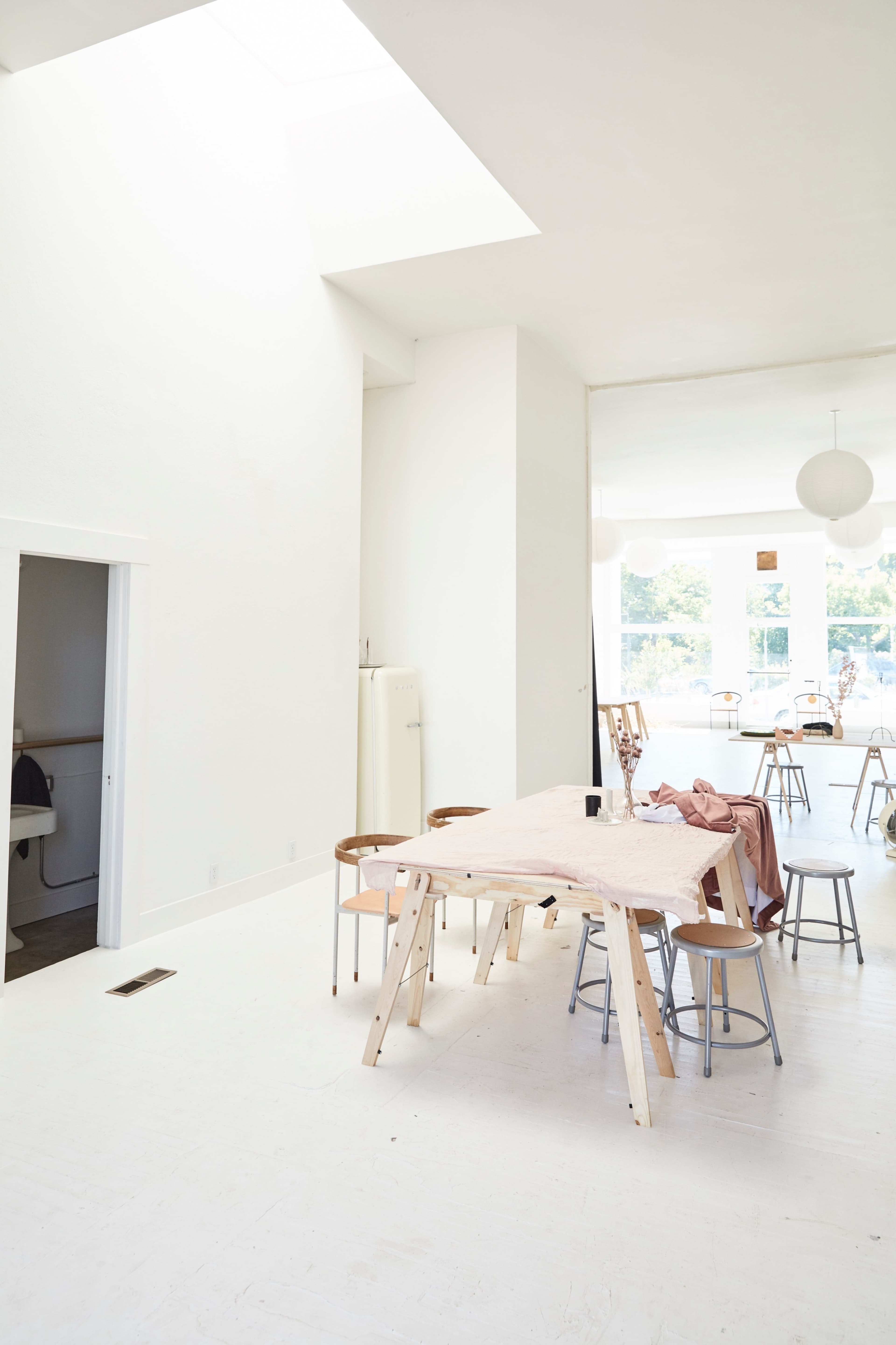 A bright, minimalist space features a central wooden table surrounded by chairs, with large windows allowing natural light to fill the room.