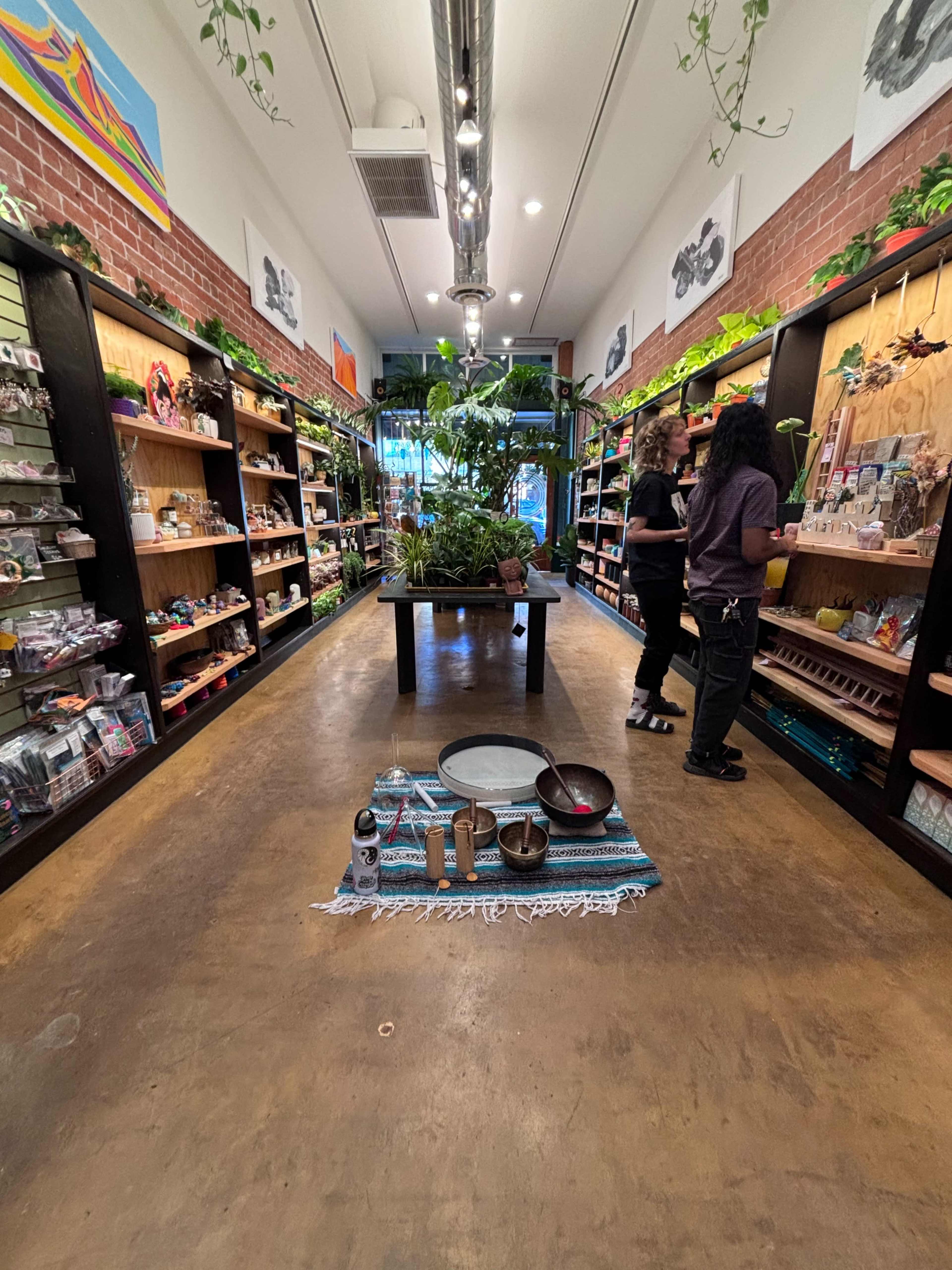The image shows a retail store with wooden shelves filled with various products on either side, and customers browsing in the center aisle.