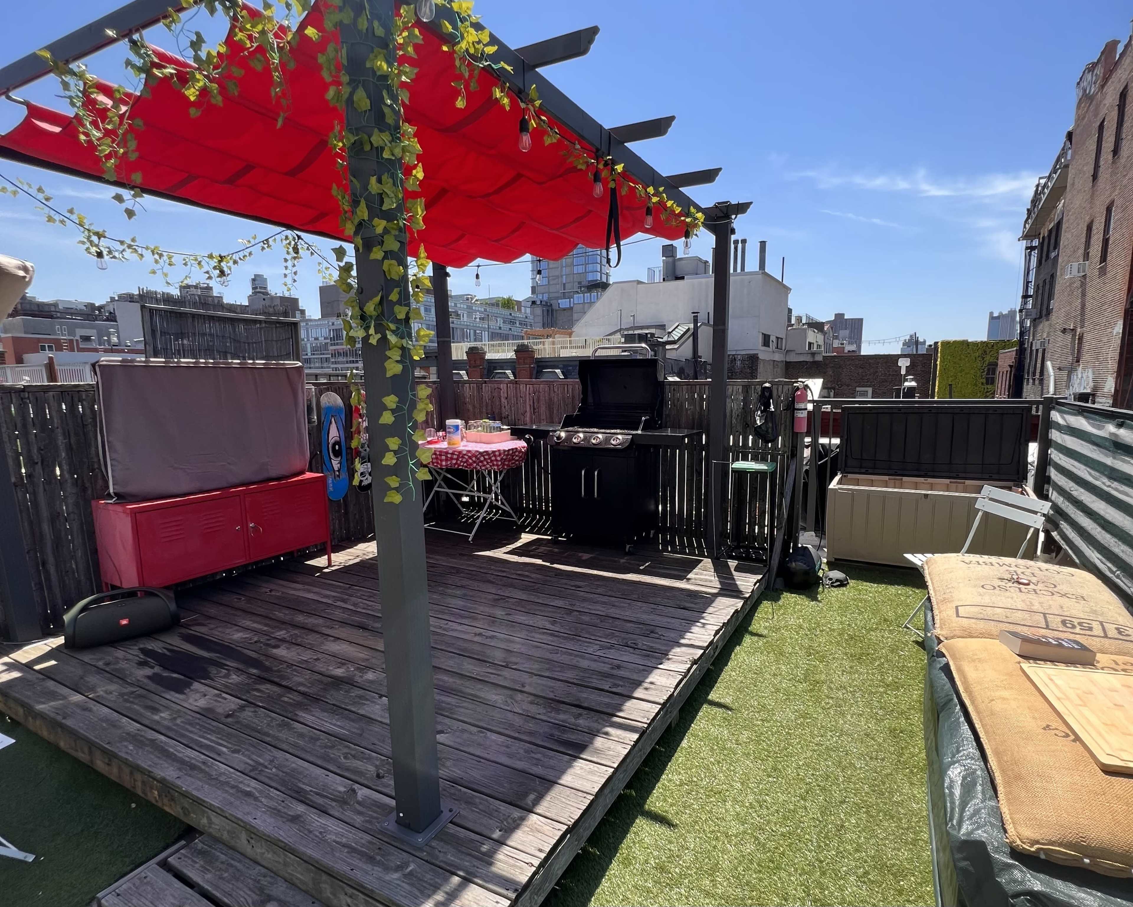 The image shows a rooftop patio with a red canopy, a barbecue grill, a table with pink and white tablecloth, and a view of surrounding buildings under a clear blue sky.