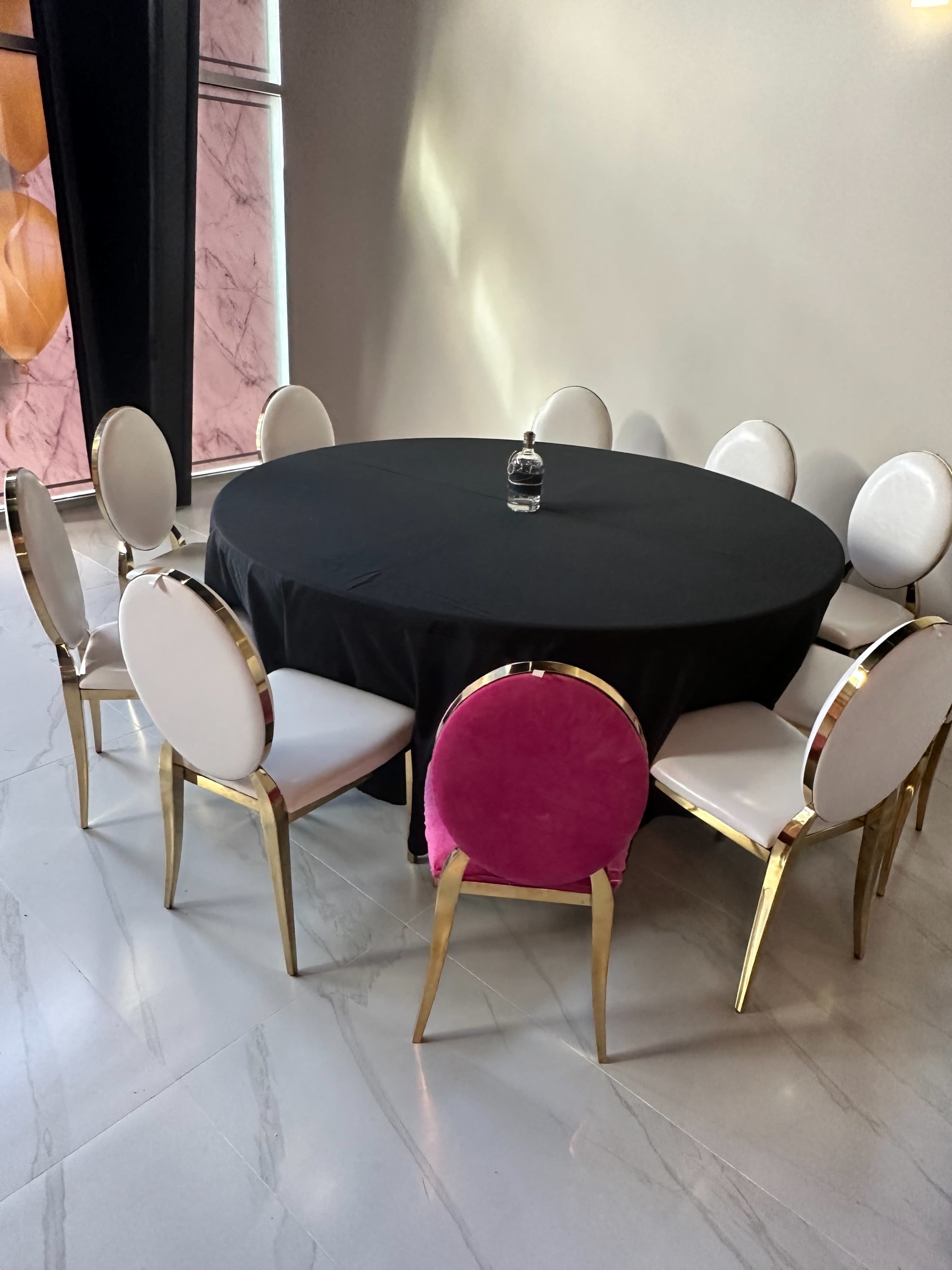 A round table covered with a black tablecloth is surrounded by ten chairs, five with white seats and one with a pink seat.