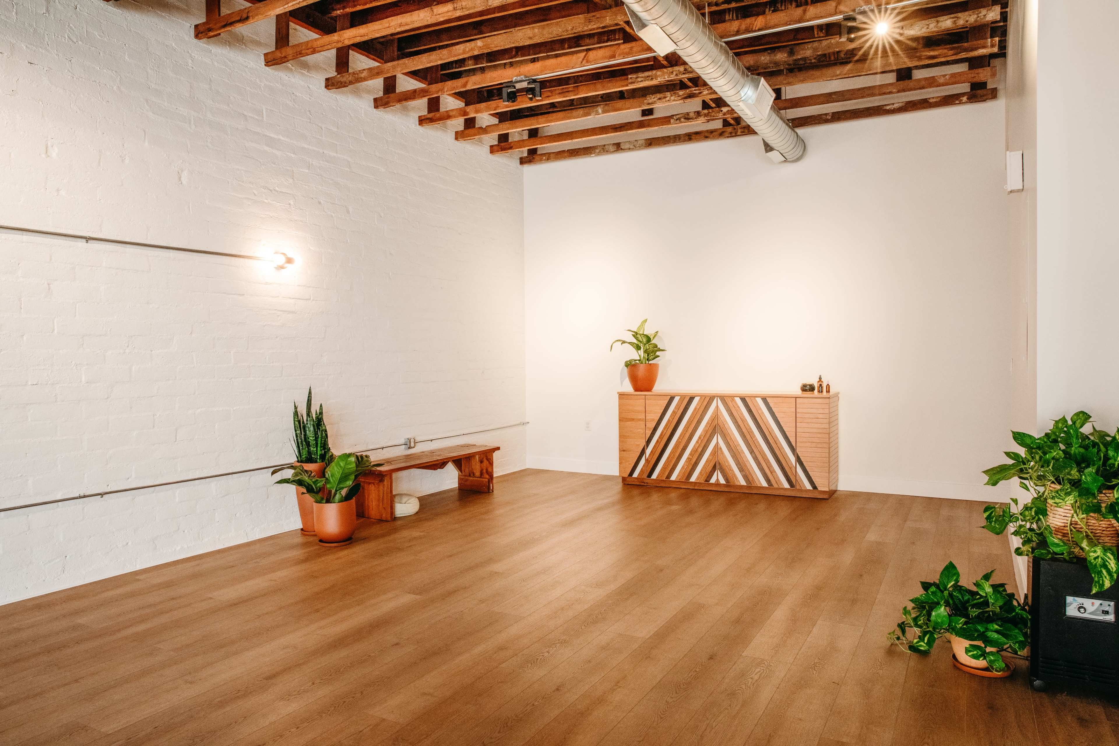 The image shows a spacious, minimalist interior with wooden beams, a light-colored wall, a wooden cabinet with a geometric design, and several potted plants arranged around a wooden bench.