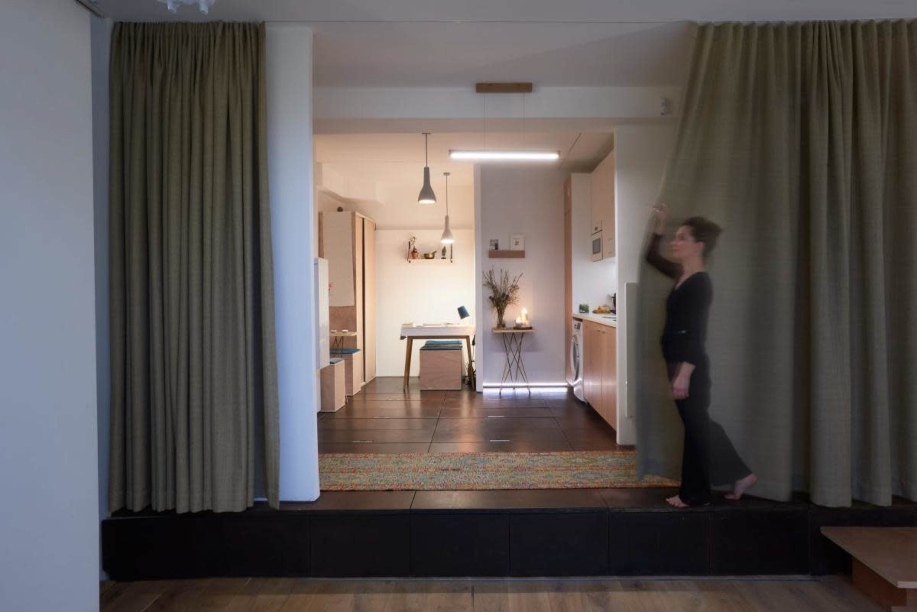 A woman walks past a curtain in a modern, open-plan living space that features a kitchenette and a dining area.