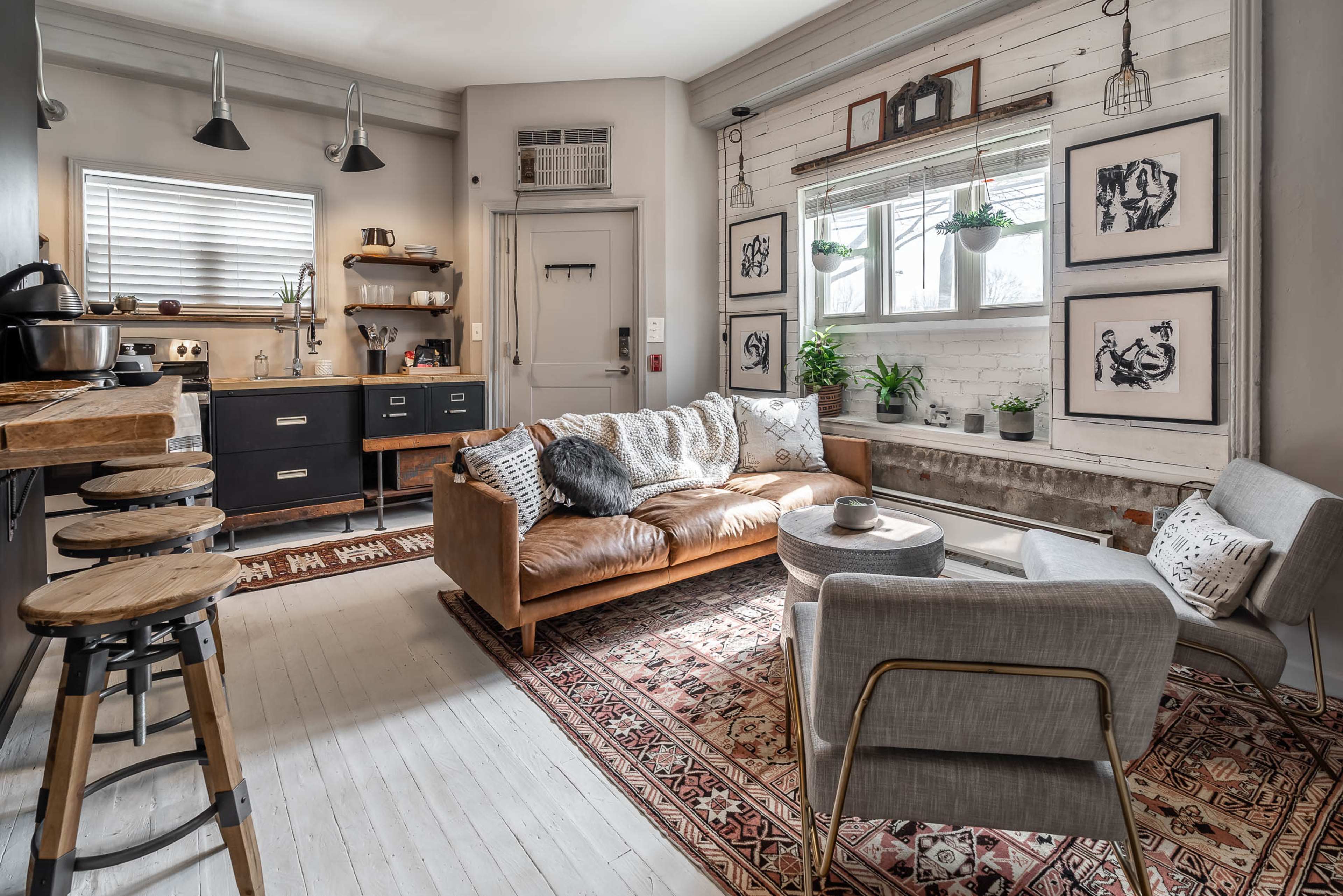 A cozy living room features a leather couch, a gray armchair, and a decorative rug, with framed art on the walls and a window allowing natural light to enter.