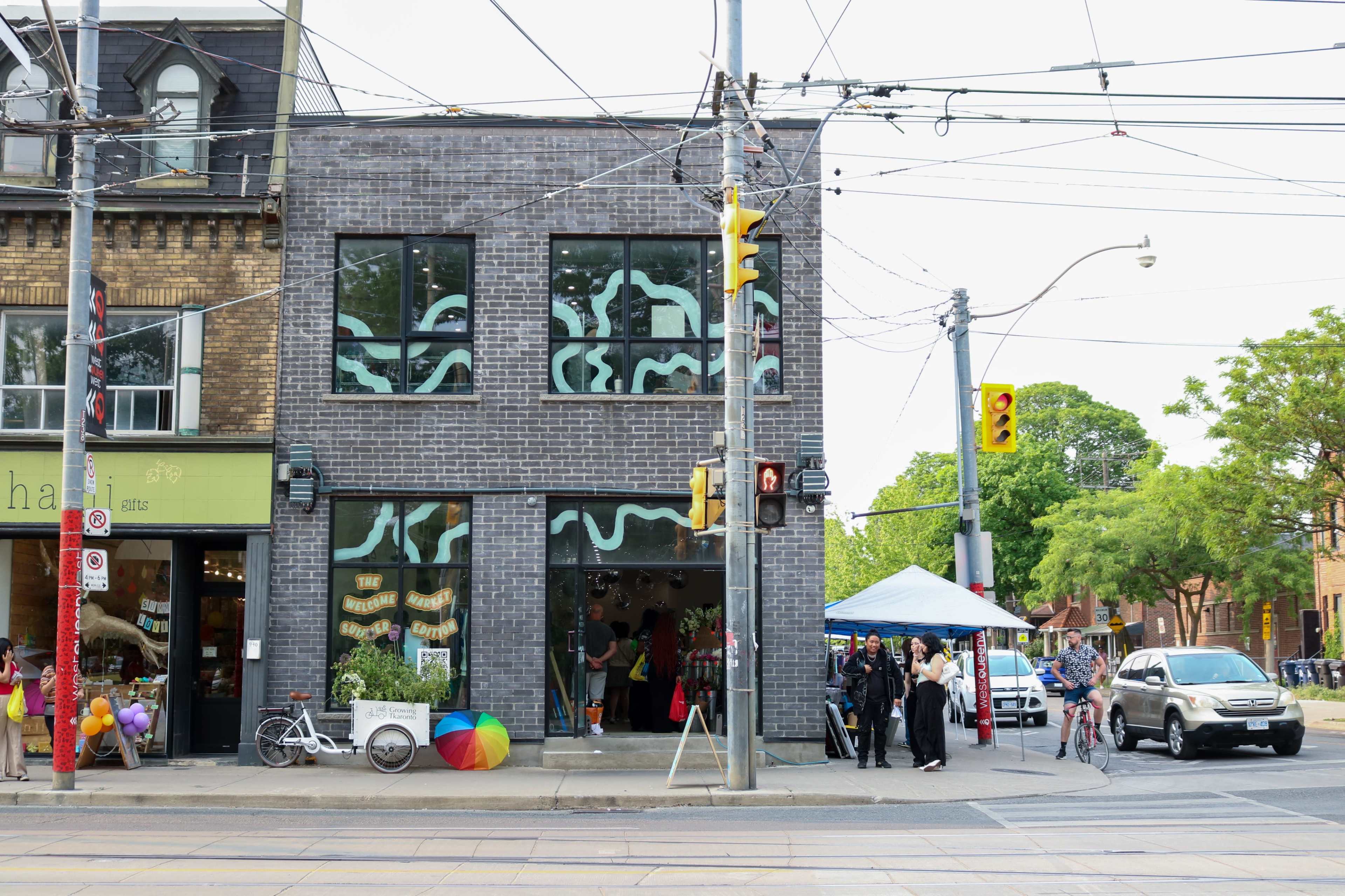 The scene features a modern brick building with large windows displaying colorful decorations, while pedestrians gather outside near a street with cars and a pop-up tent.