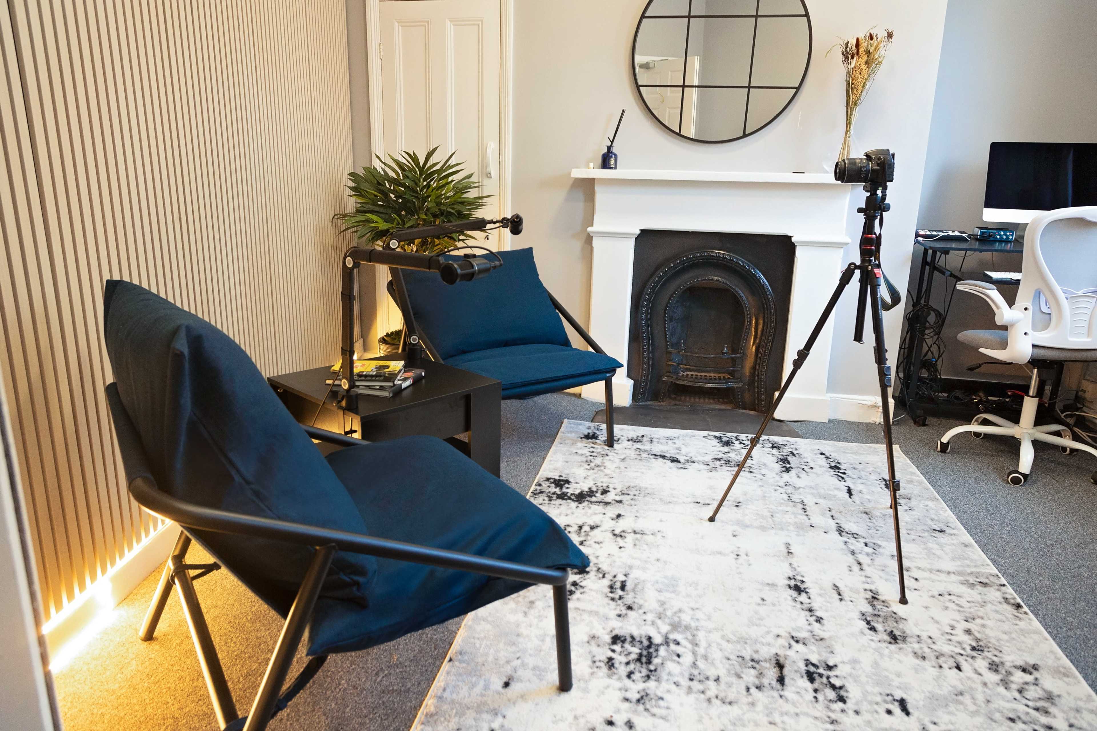 A well-lit room features two blue chairs, a small black table, a camera on a tripod, and a mirror on the wall above a decorative fireplace.