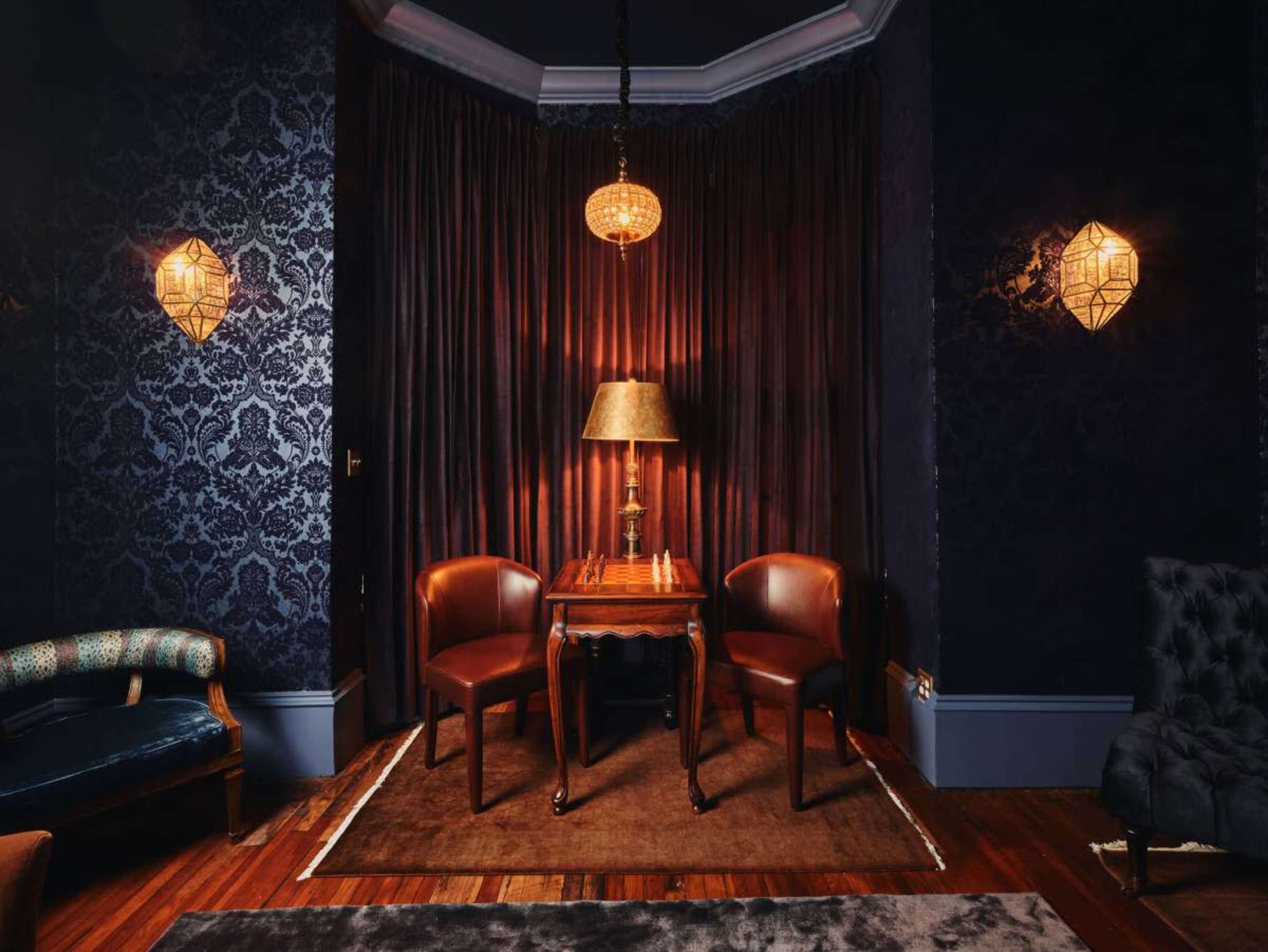 A cozy interior space features two brown chairs and a small wooden table, illuminated by a pendant lamp and framed by dark, patterned wallpaper and curtains.