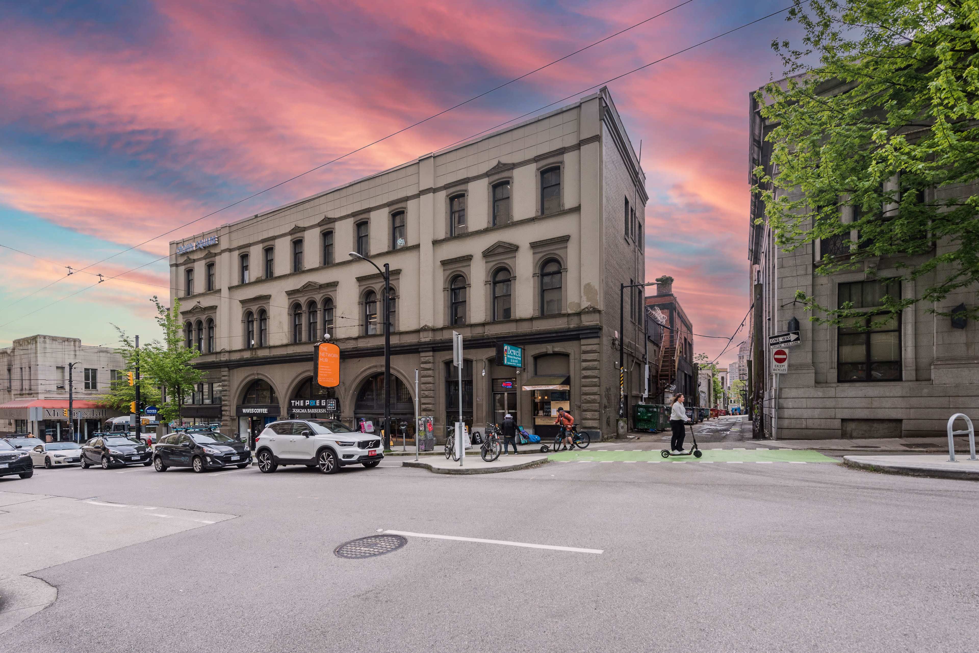 A historic building sits at the intersection of two streets, with vehicles and pedestrians navigating the area under a colorful sky.