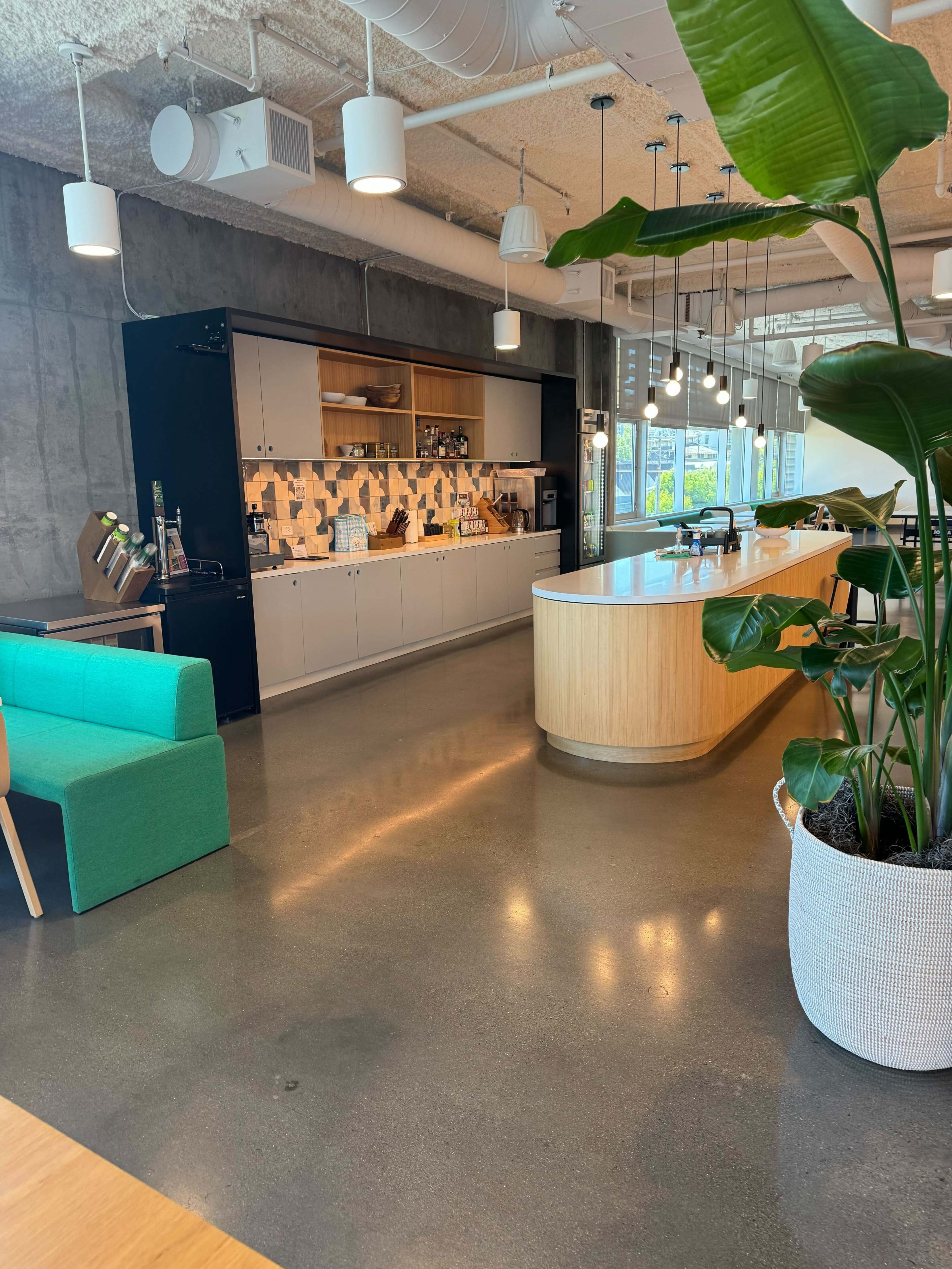 A modern office space with a teal sofa, a kitchen area featuring a counter and decorative tiles, and large windows allowing natural light.