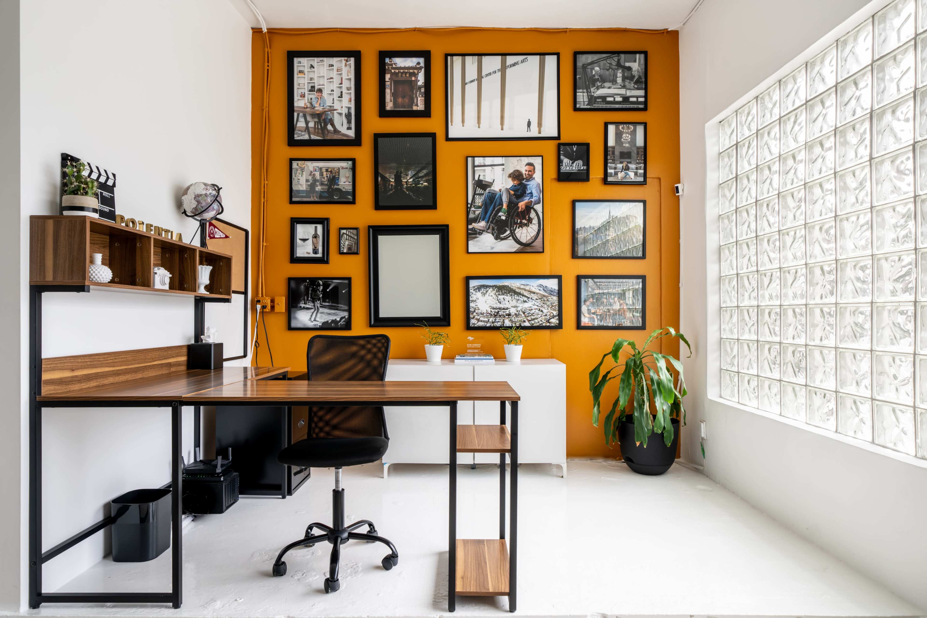A modern home office features a wooden desk and a black chair facing a gallery wall of framed art against an orange backdrop.