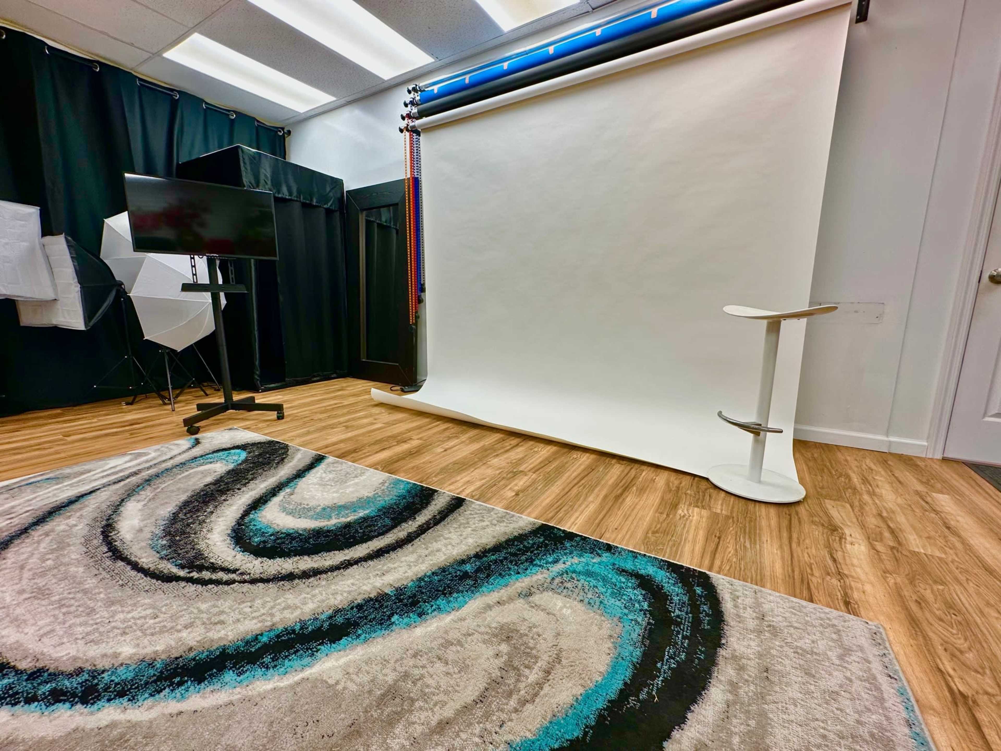 The image shows a photography studio equipped with a backdrop setup, a monitor on a stand, and a patterned rug on the floor.