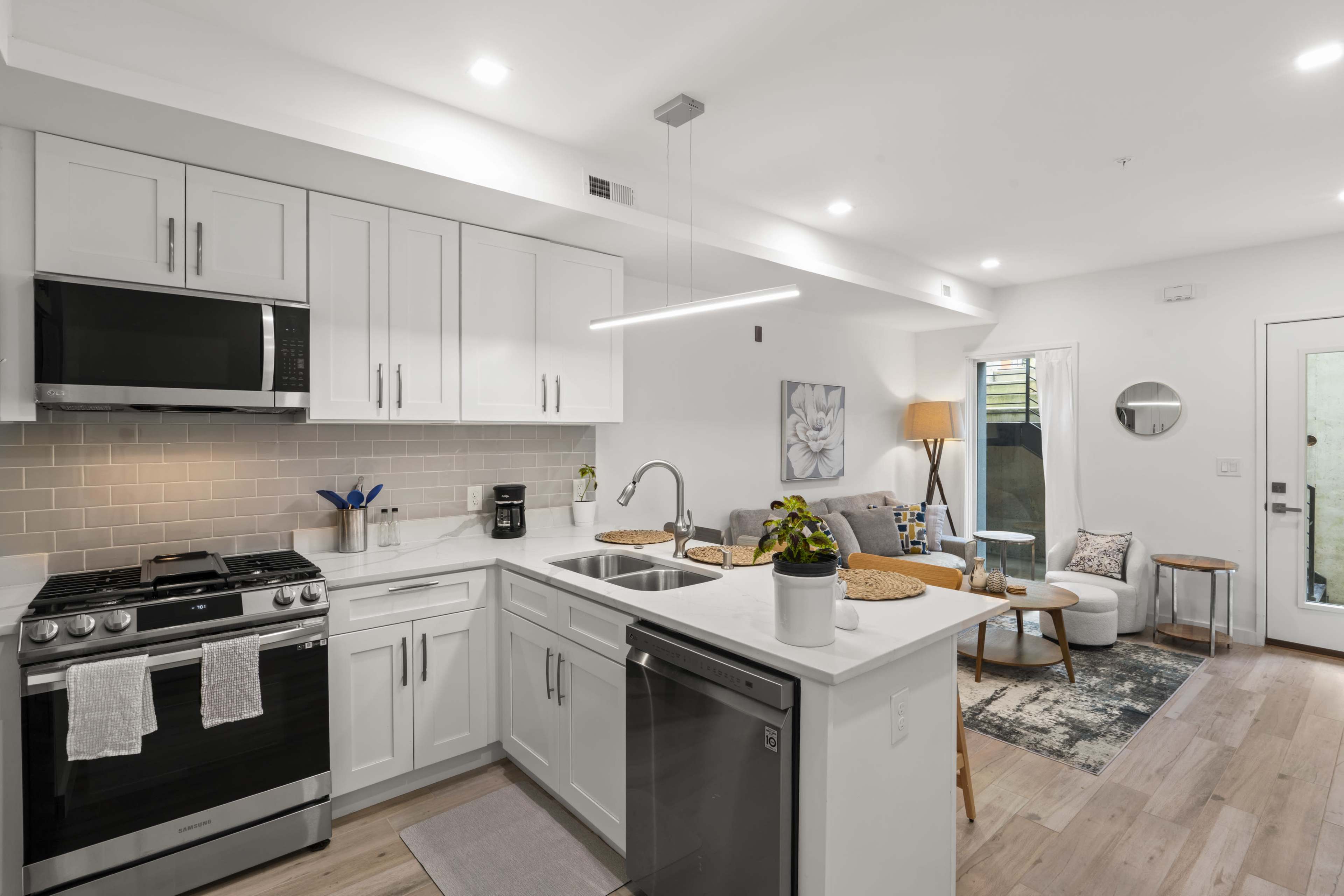 The image shows a modern kitchen featuring white cabinets, stainless steel appliances, and an open layout connecting to a living area with a sofa and a table.