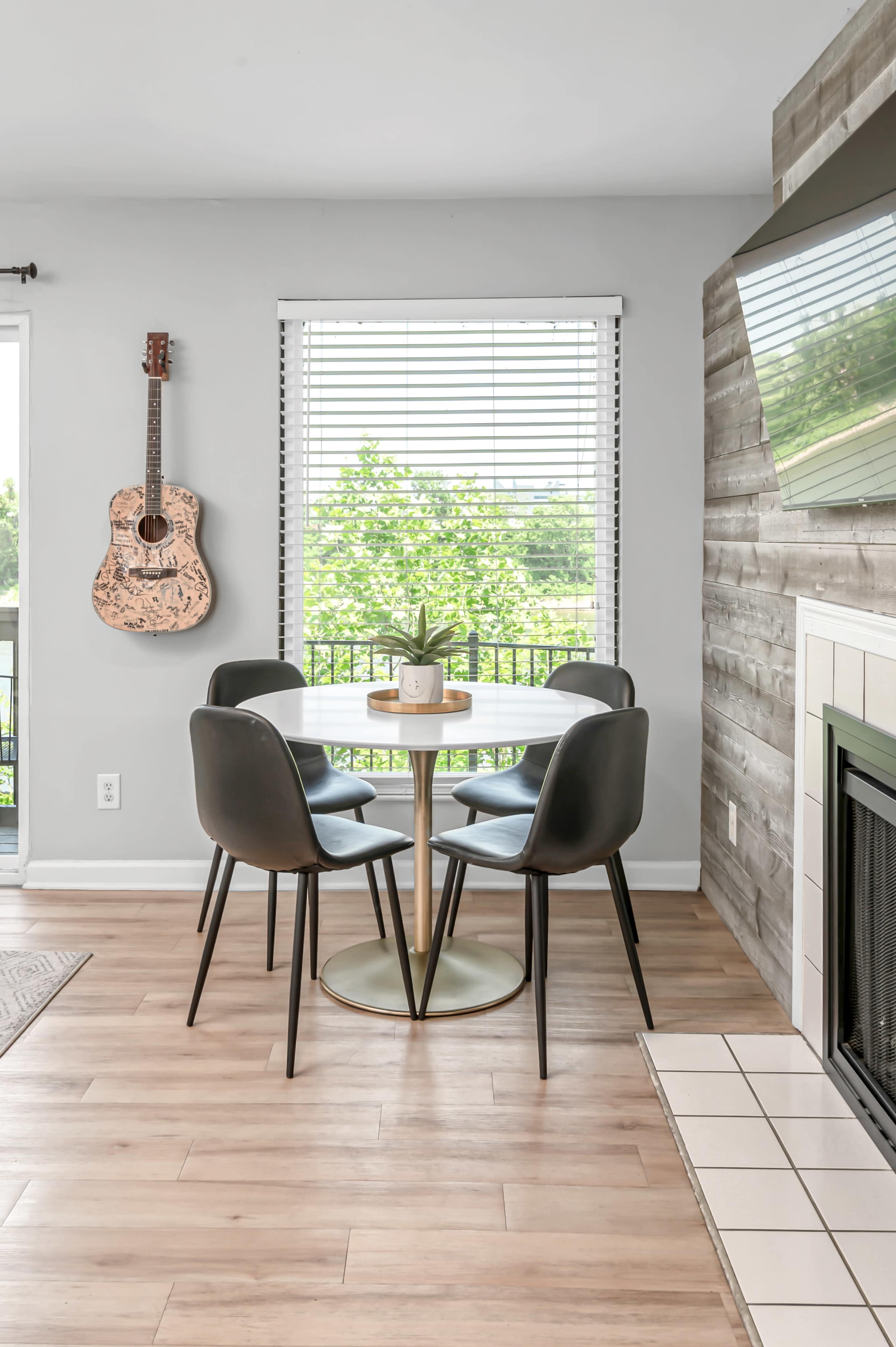 A circular dining table with four black chairs is positioned near a window with blinds, overlooking greenery, and a guitar hangs on the wall beside a fireplace.