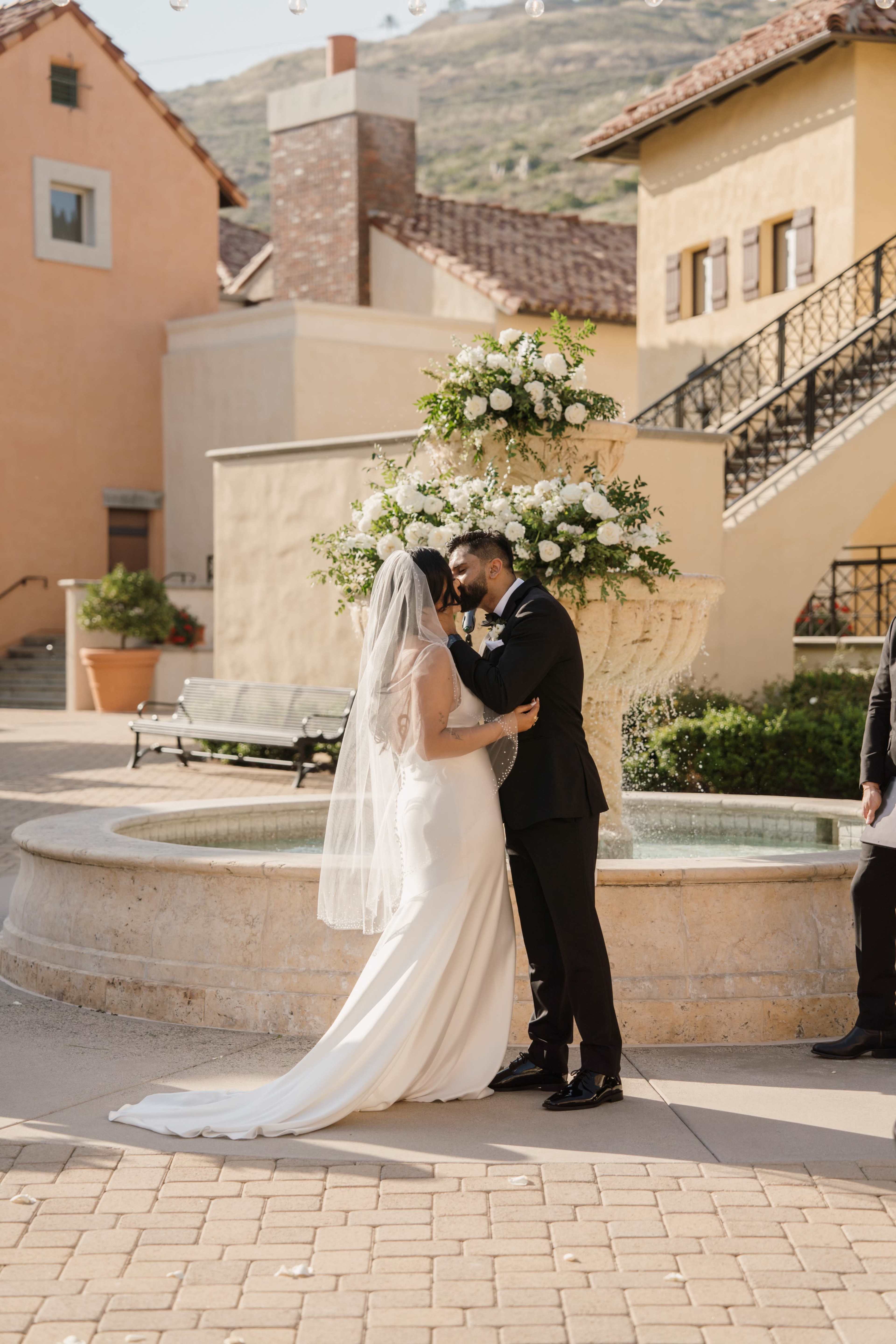 A couple in formal attire embraces in front of a decorated fountain in a picturesque outdoor setting.