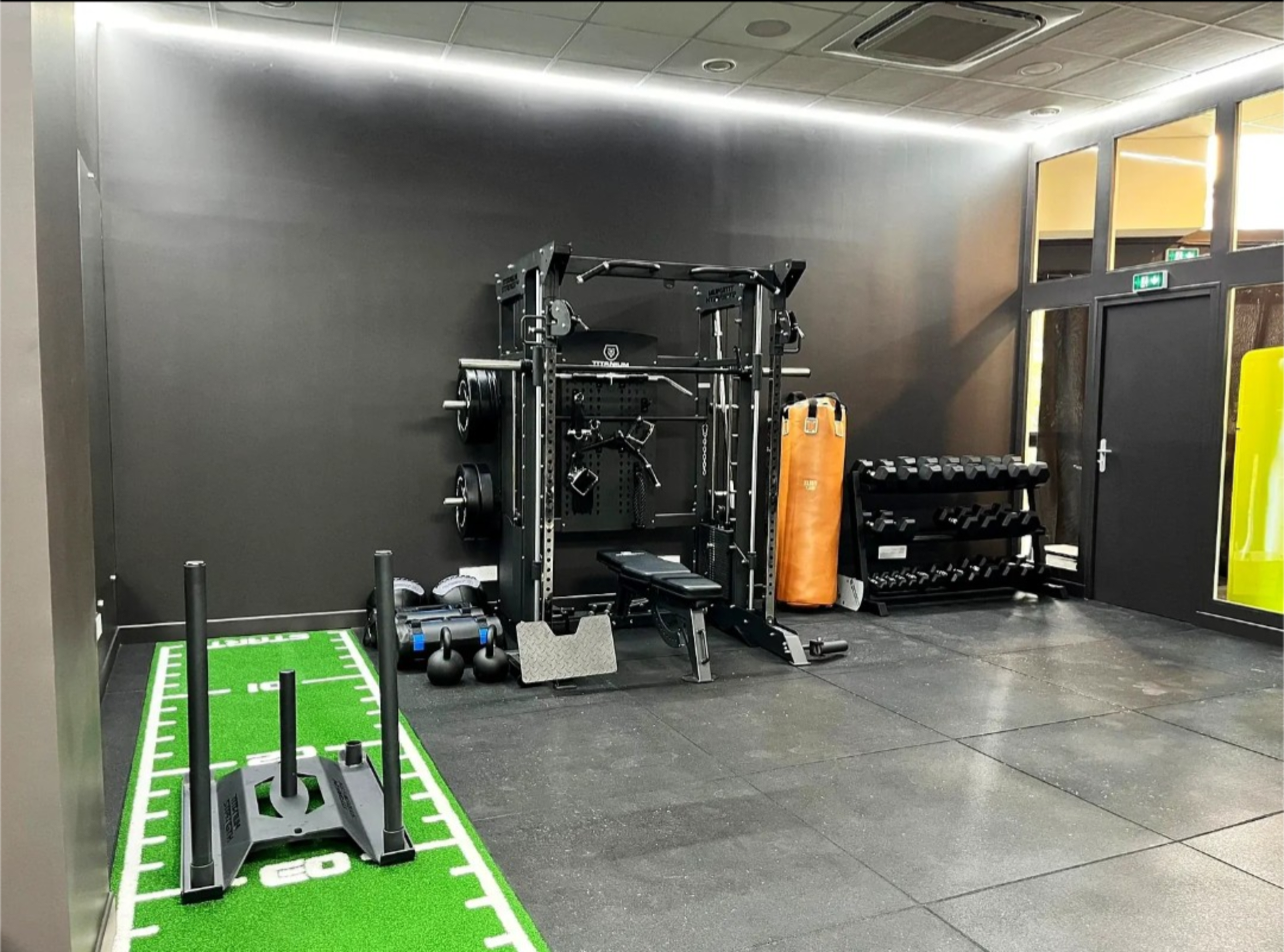The image shows a modern gym space featuring a weightlifting station, a sled push area, an orange punching bag, and various dumbbells arranged on the floor.