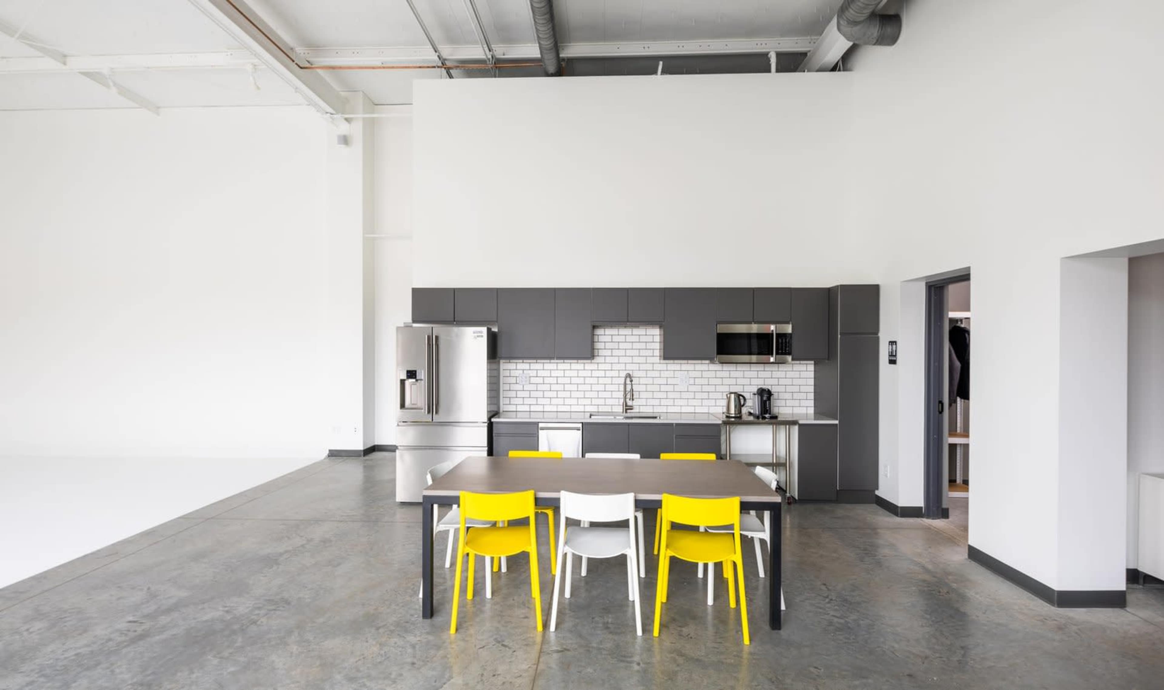 The image shows a modern kitchen area with a gray cabinetry, stainless steel appliances, and a table surrounded by yellow and white chairs.
