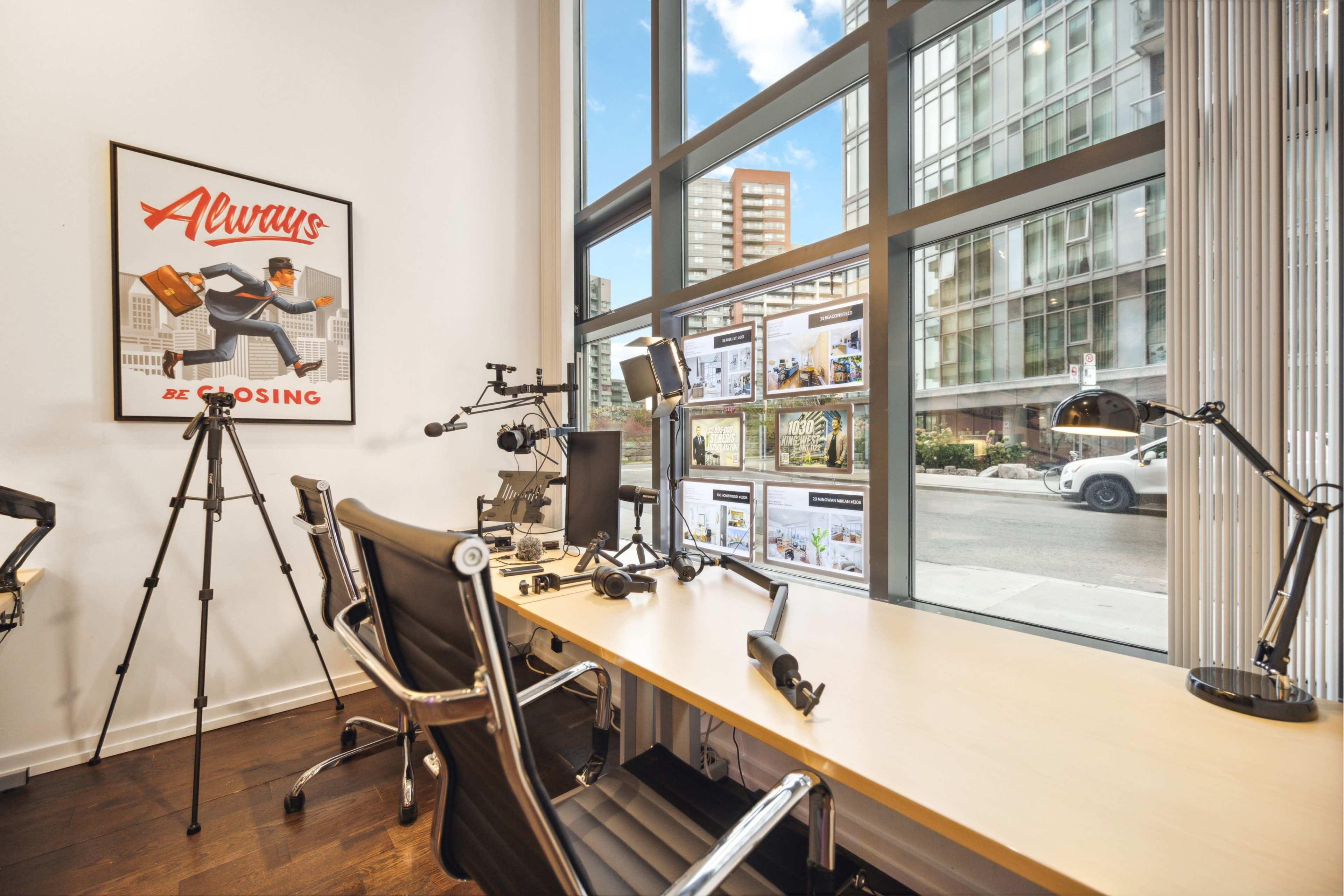 The image shows a modern office space with a large window, a desk equipped with multiple monitors, and various tripods and desk lamps.