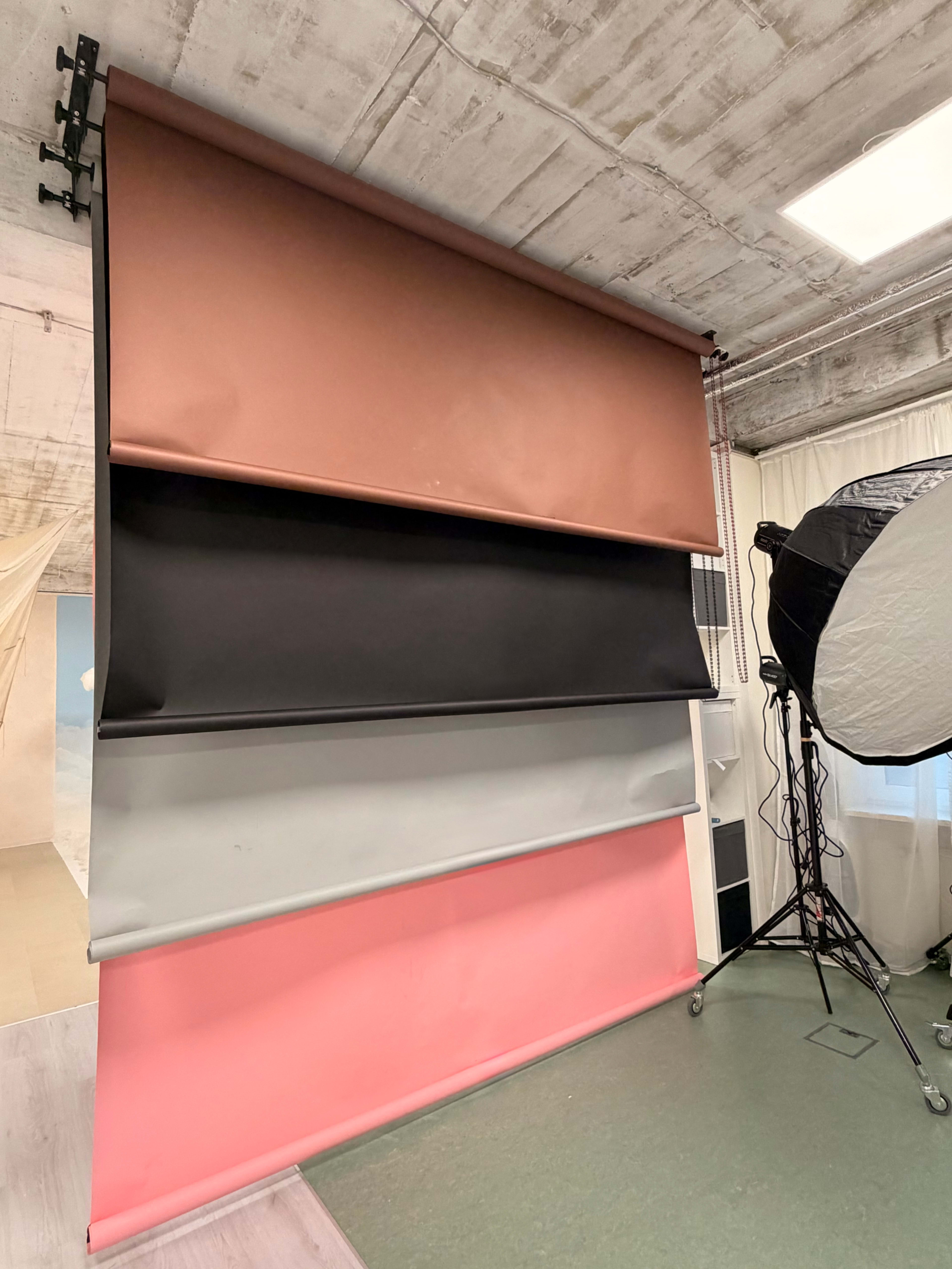 The image shows a photography studio backdrop featuring four rolled fabric panels in colors of pink, gray, black, and brown, arranged vertically on a wall.