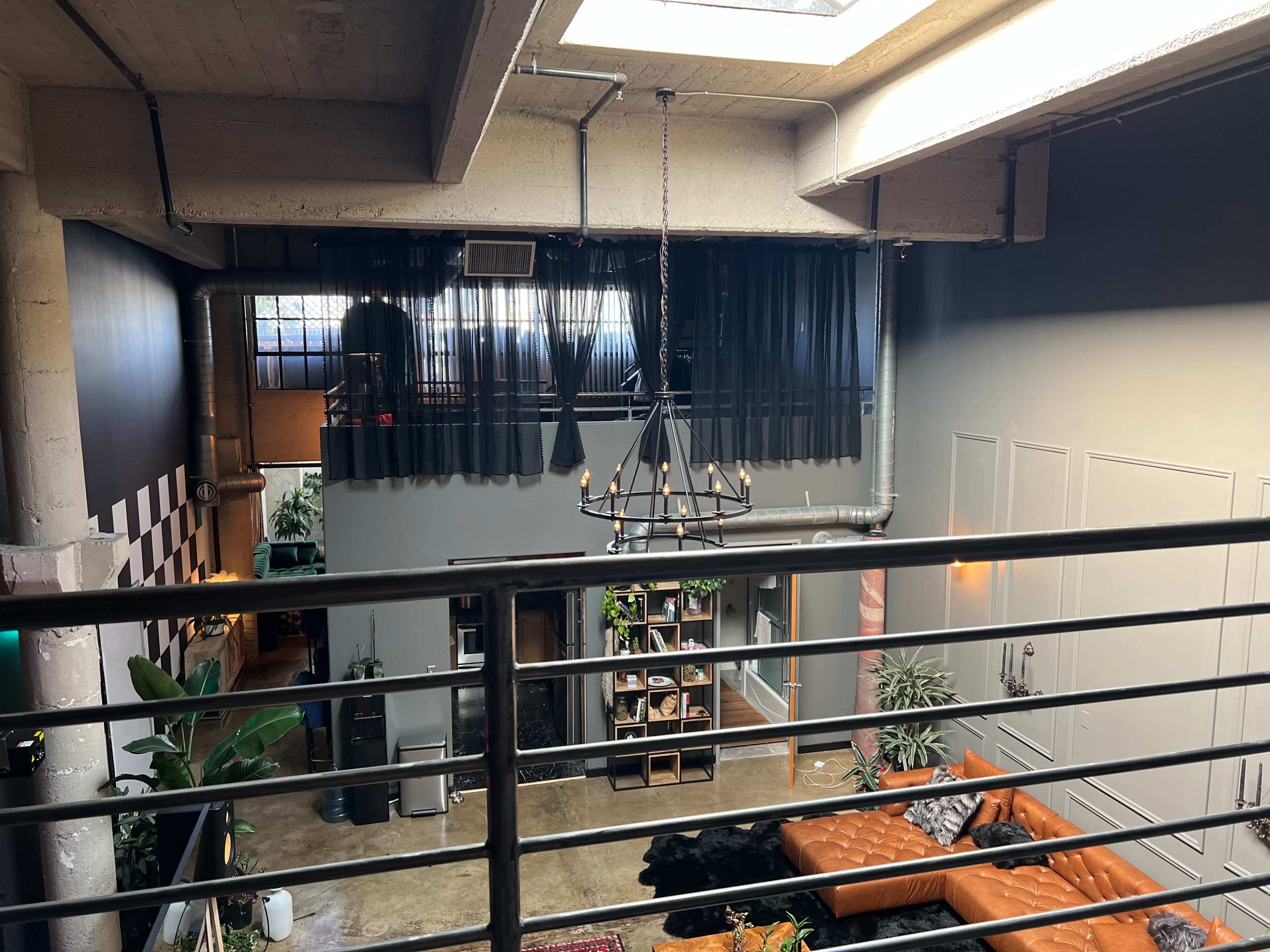 A spacious loft with a second-level railing overlooking a living area furnished with a leather sofa and decorated with plants, while large windows provide natural light.