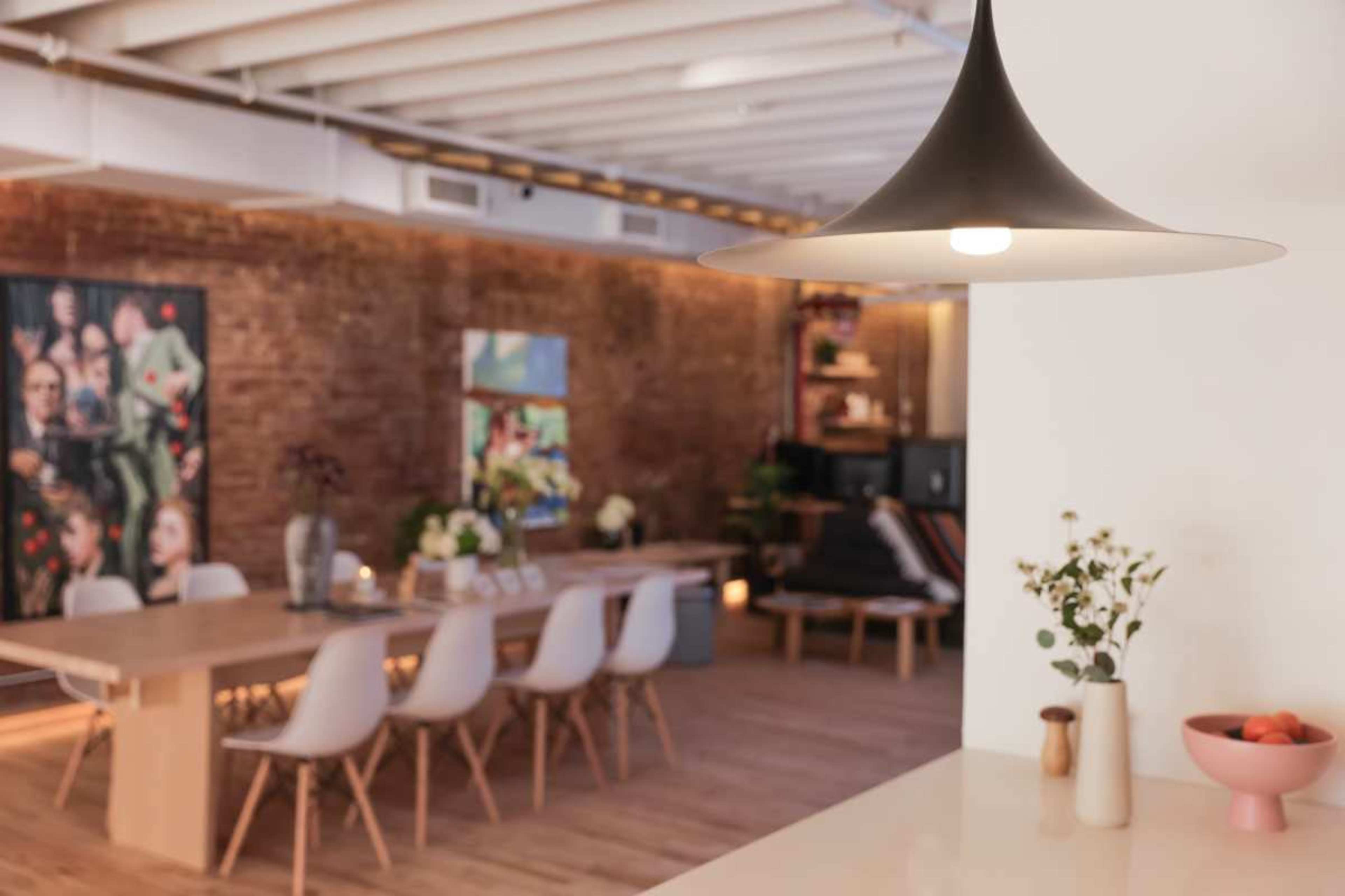 A modern dining area features a long wooden table surrounded by white chairs, with exposed brick walls and a large pendant light overhead.