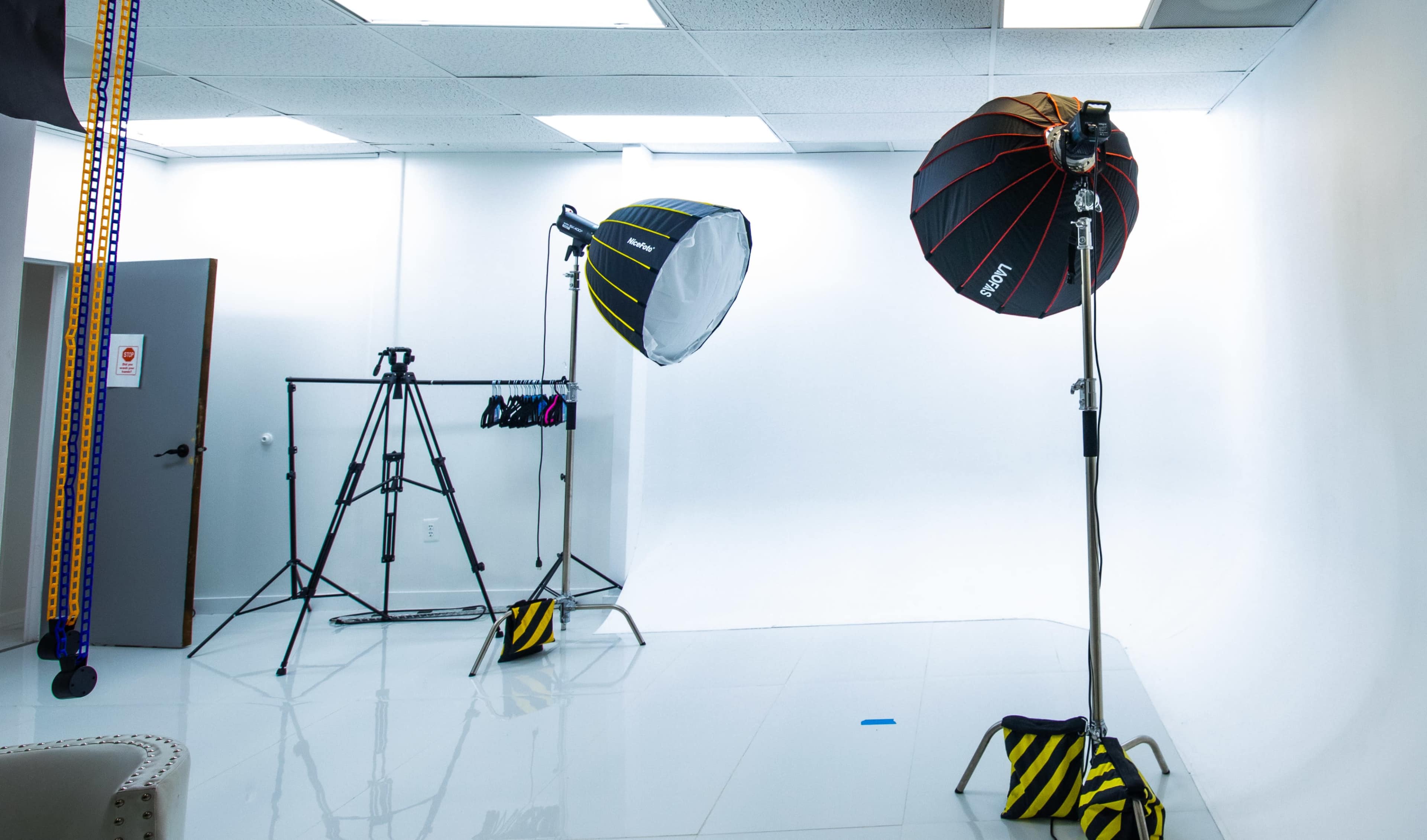 A photography studio with two light stands, a tripod, and a blank white backdrop.