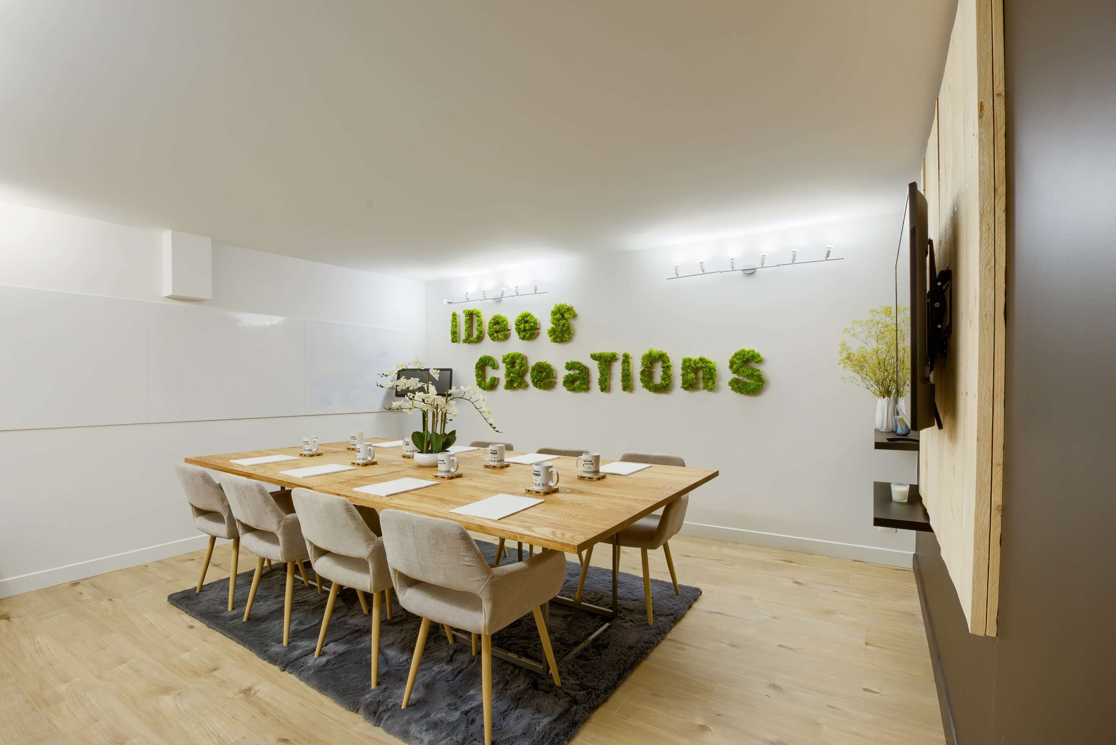 A modern conference room features a wooden table surrounded by beige chairs, with green moss spelling out "Ideas & Creations" on the wall.