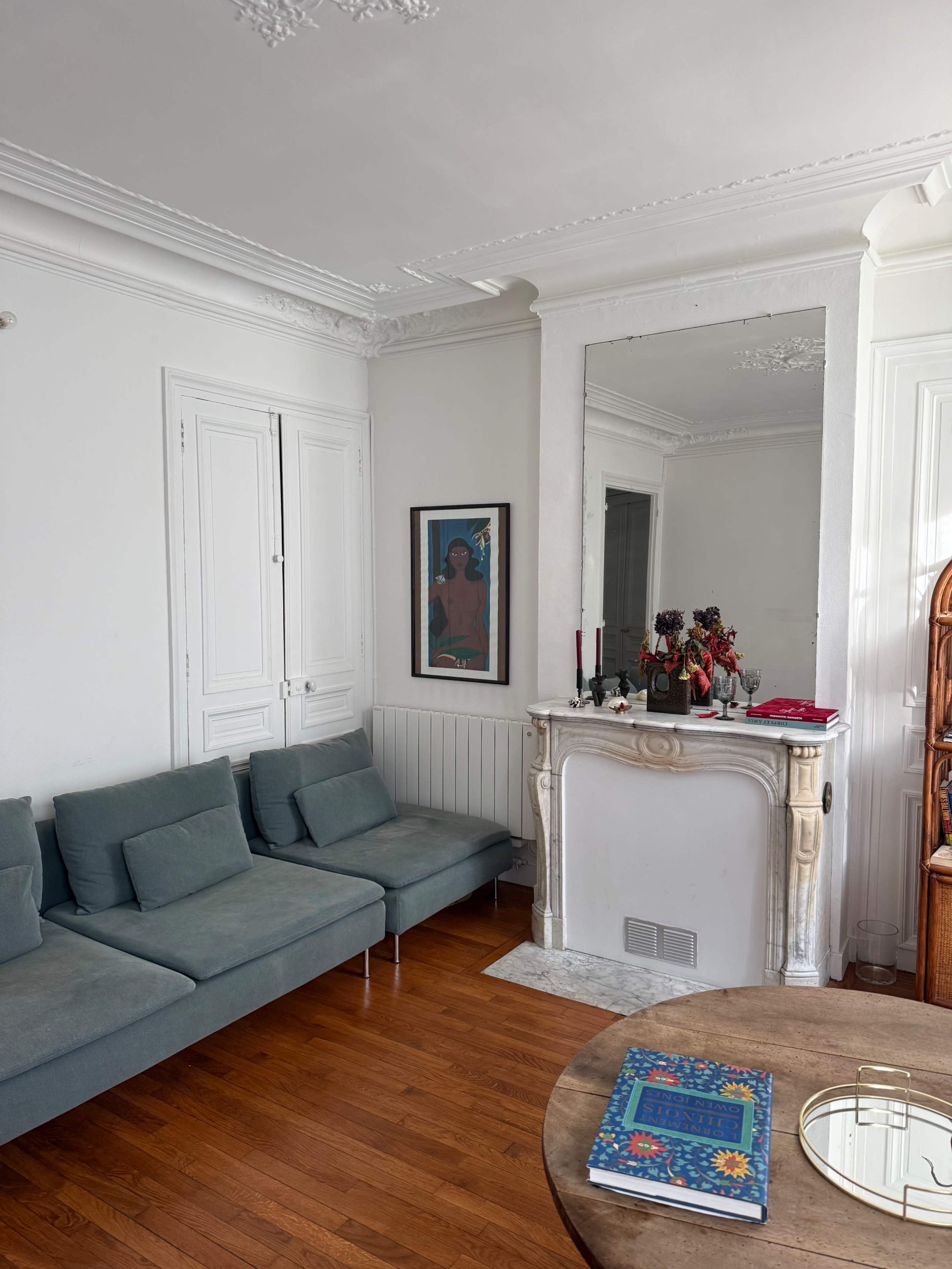 The image shows a living room with a green sectional sofa, a wooden table, a decorative mirror, and a mantelpiece featuring small statues and candles.