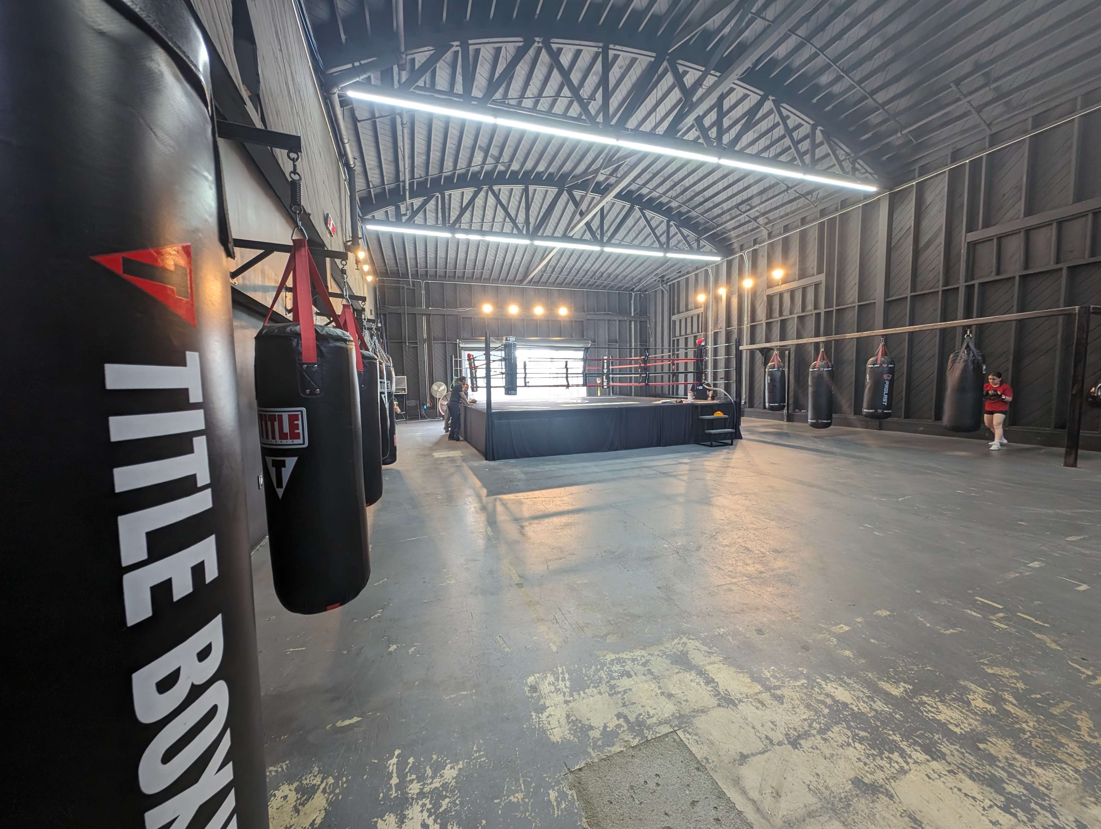 A spacious boxing gym featuring a central boxing ring surrounded by heavy punching bags and bright overhead lights.