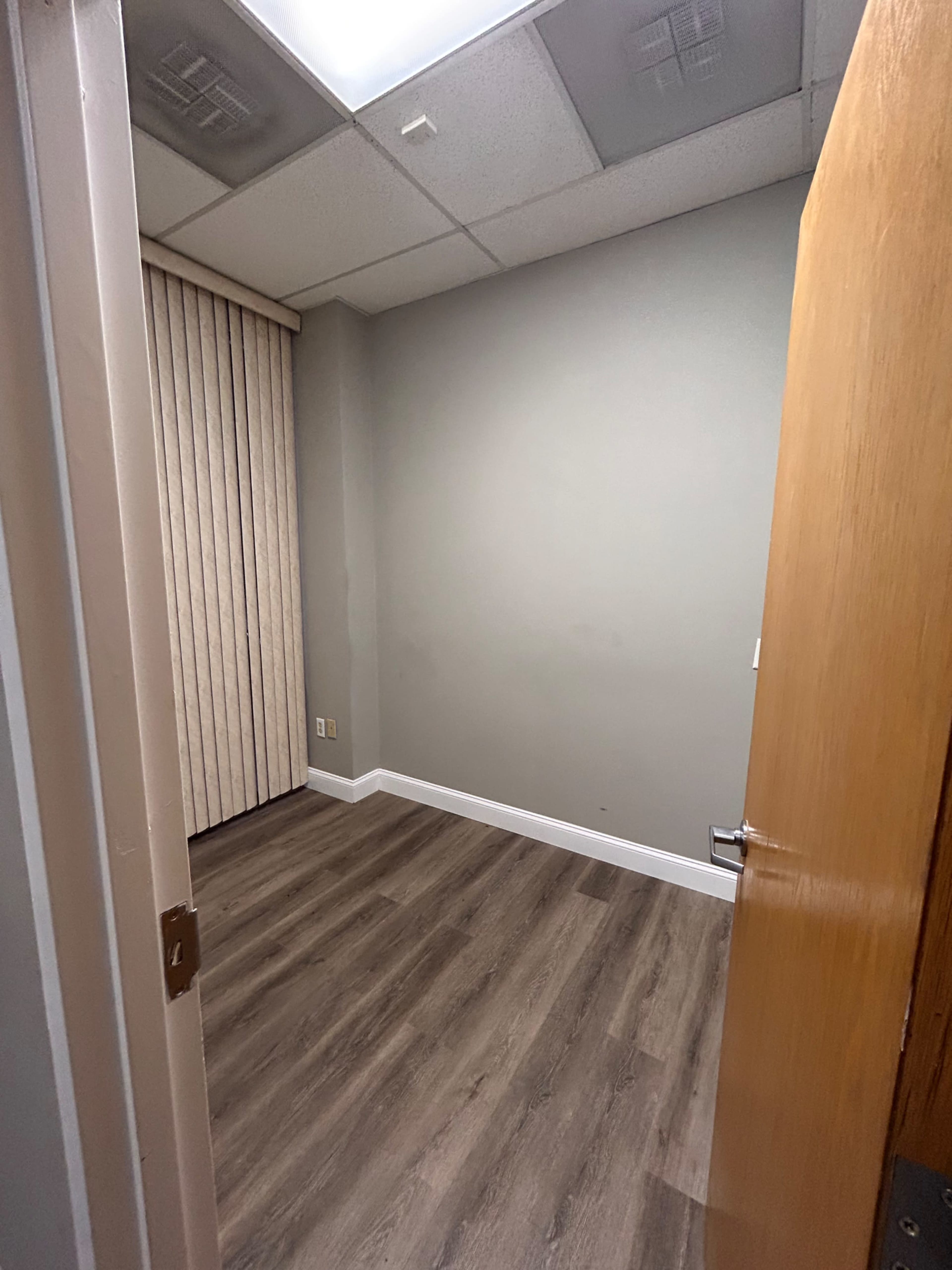 A small, empty room with light gray walls, a wooden door partially open, and laminate flooring.