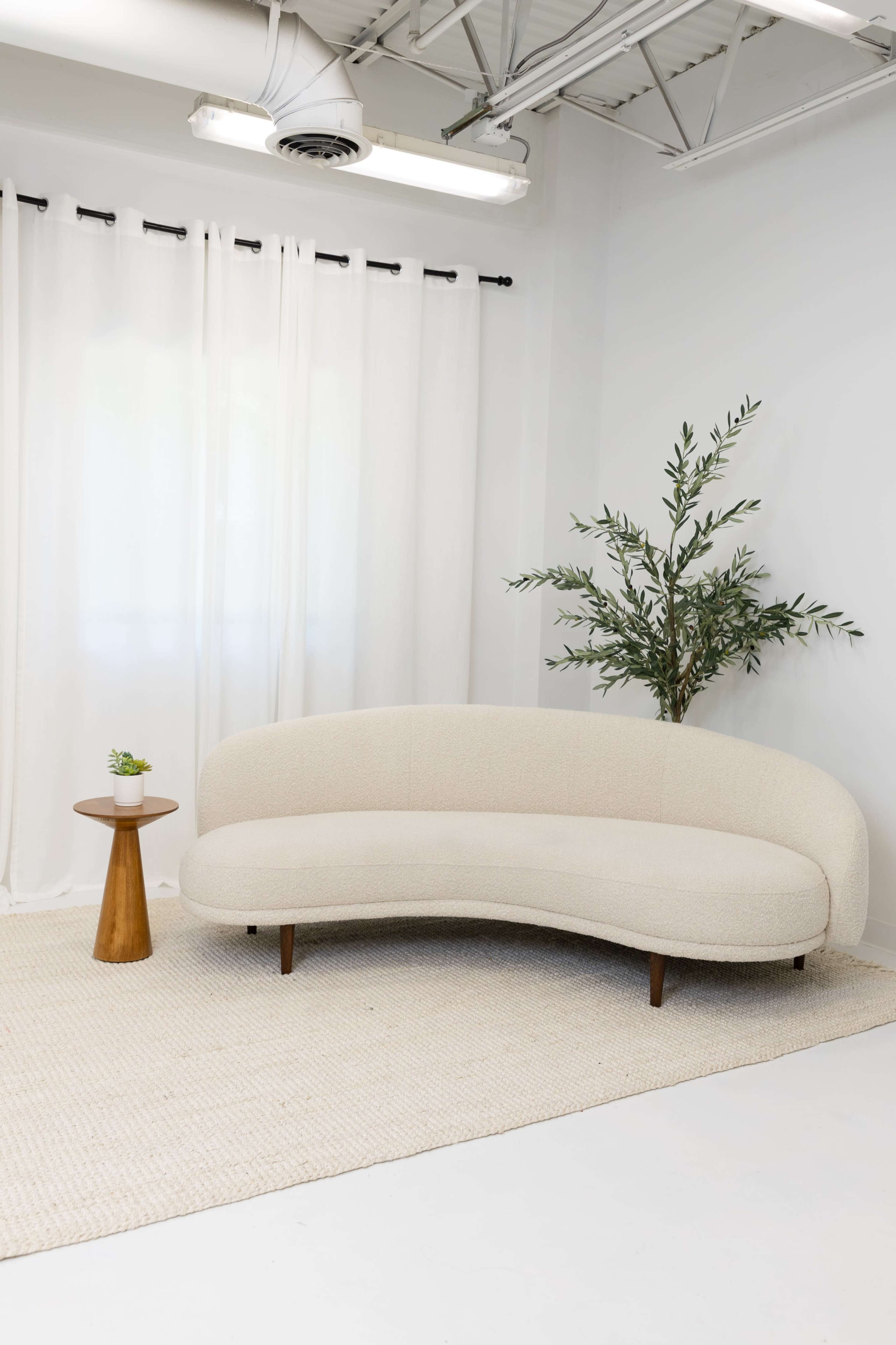 A curved beige sofa is positioned next to a small round table and a potted plant in a room with white walls and sheer curtains.