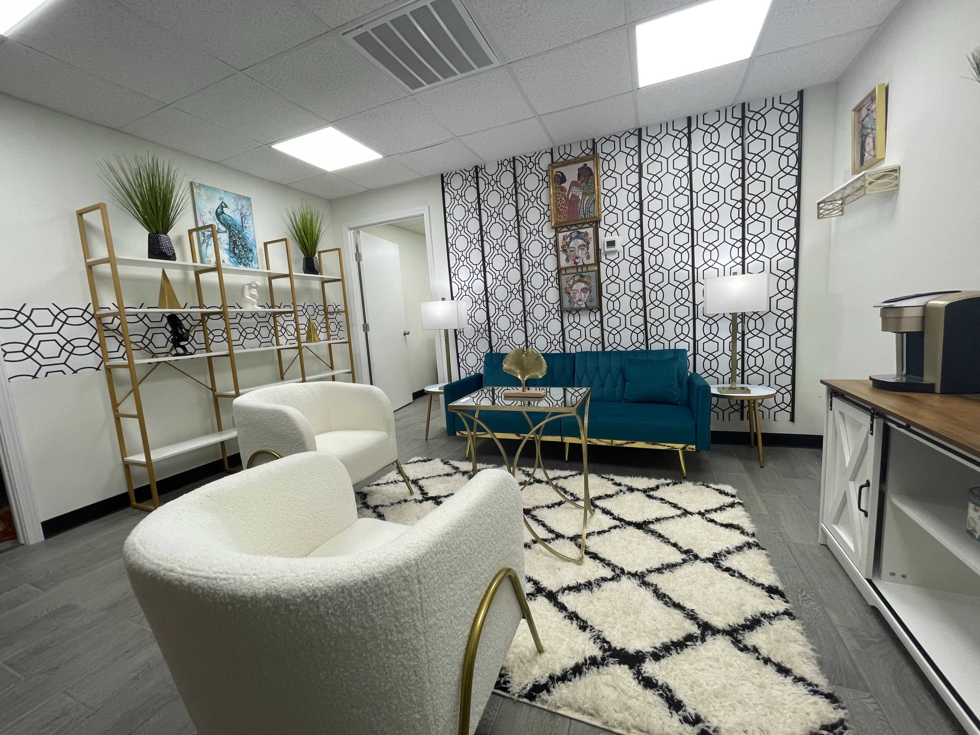 A stylish office waiting area featuring a teal couch, two white armchairs, a coffee table, and decorative shelving against a patterned wall.