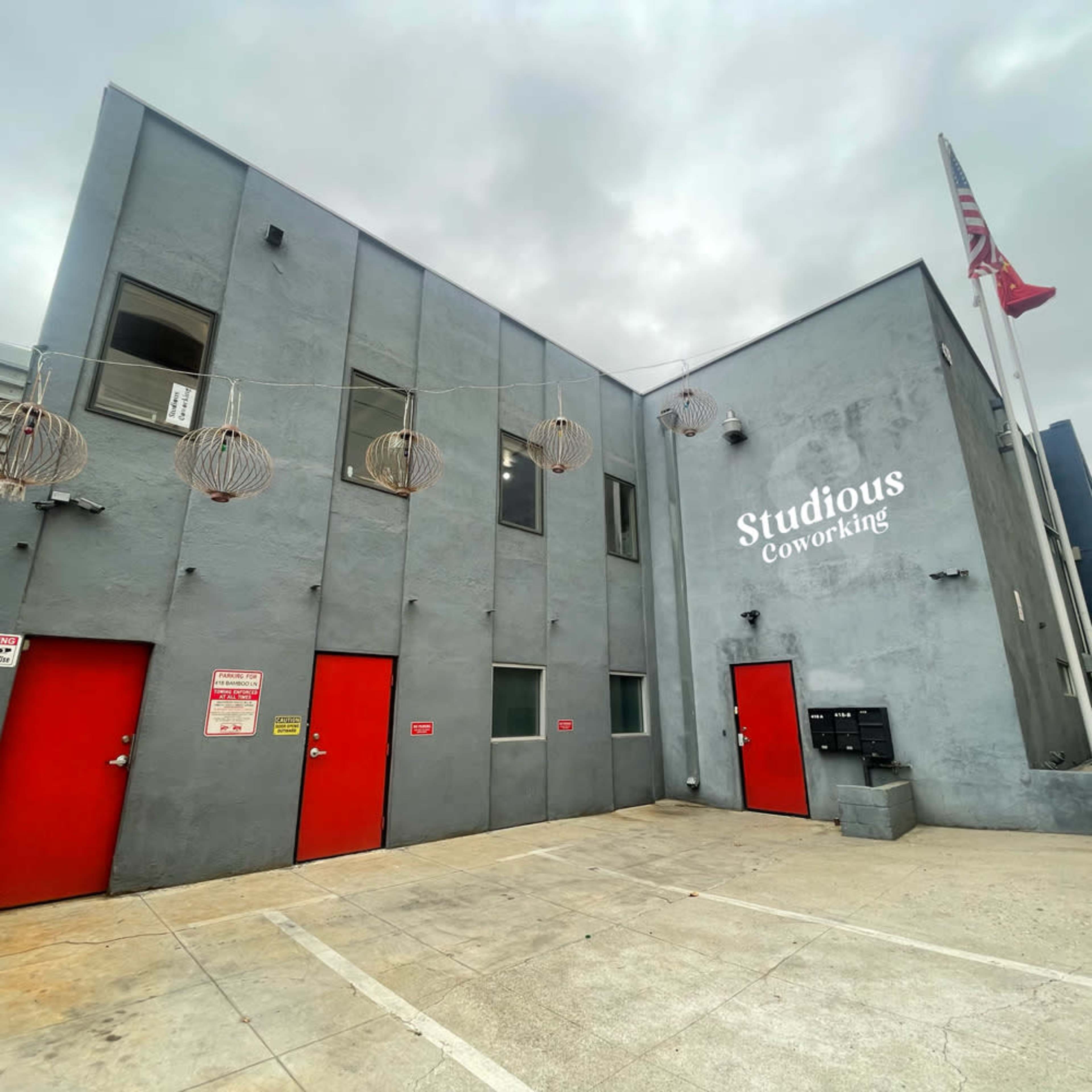 The image shows a gray building with red doors labeled "Studious Coworking," featuring hanging light fixtures and an American flag.