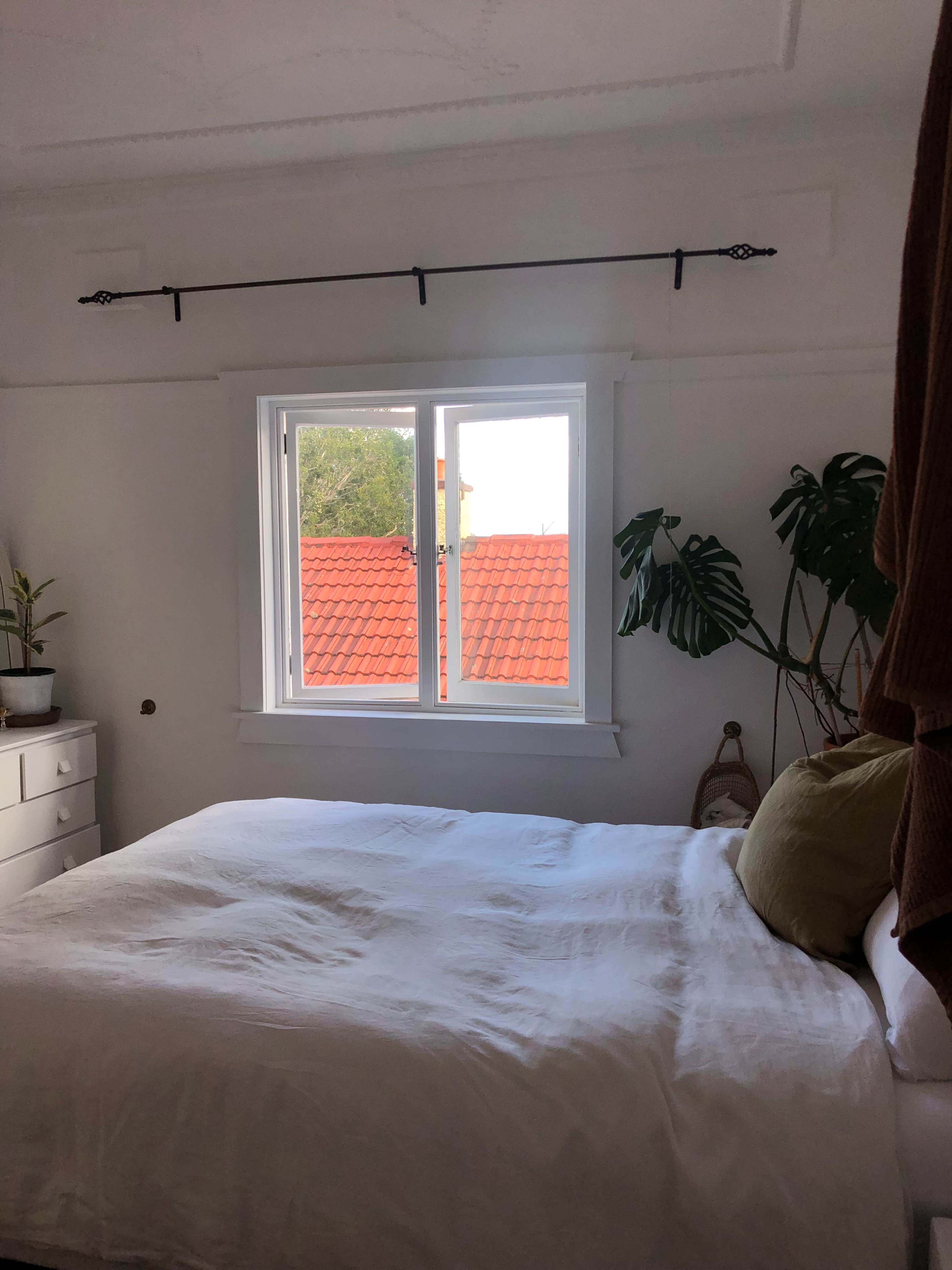 A neatly made bed with a green decorative pillow is positioned near a window that has a view of red-tiled roofs and potted plants in the room.