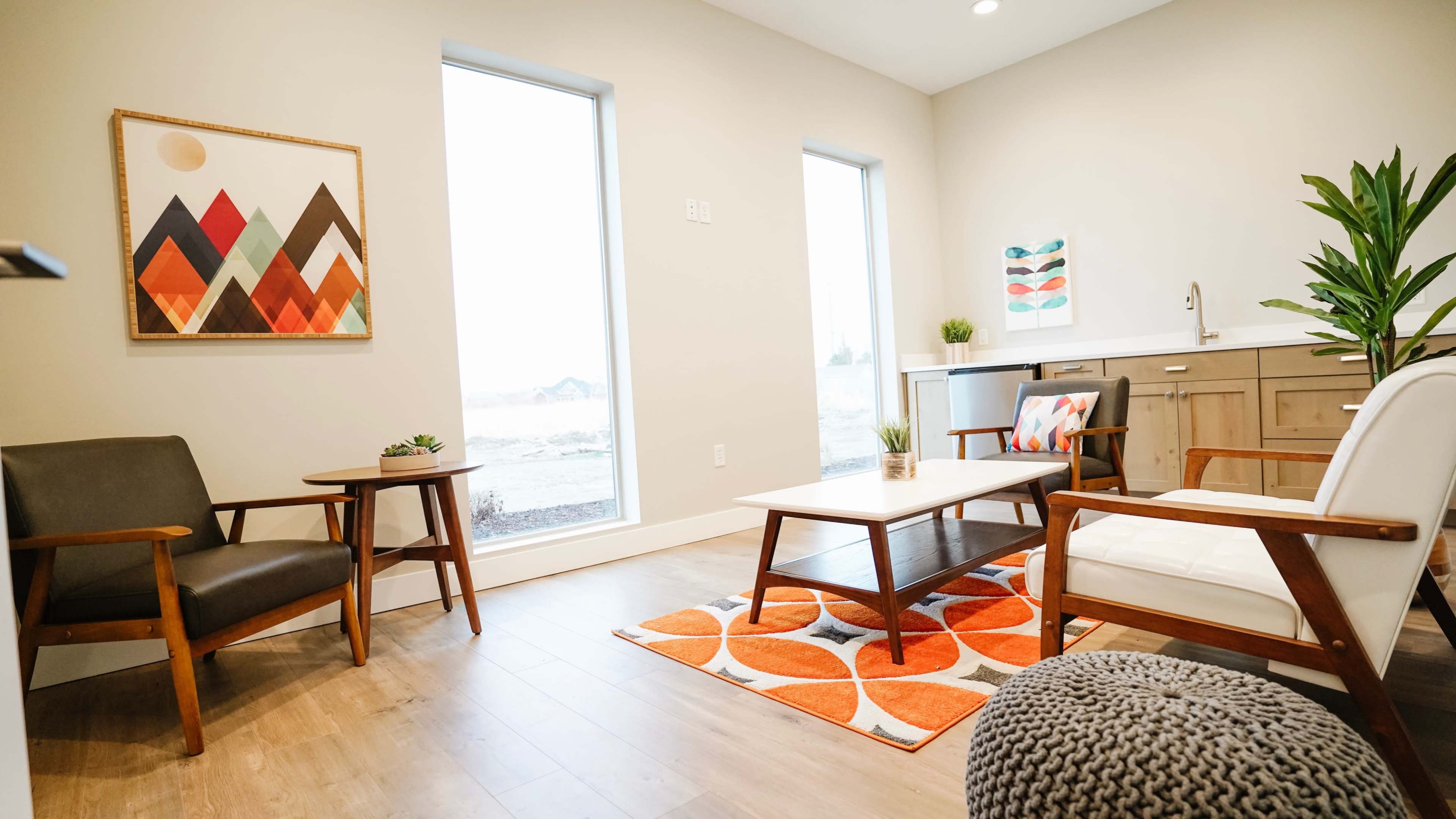 The room features two seating chairs, a coffee table, and a patterned rug, with large windows allowing natural light and a modern aesthetic.