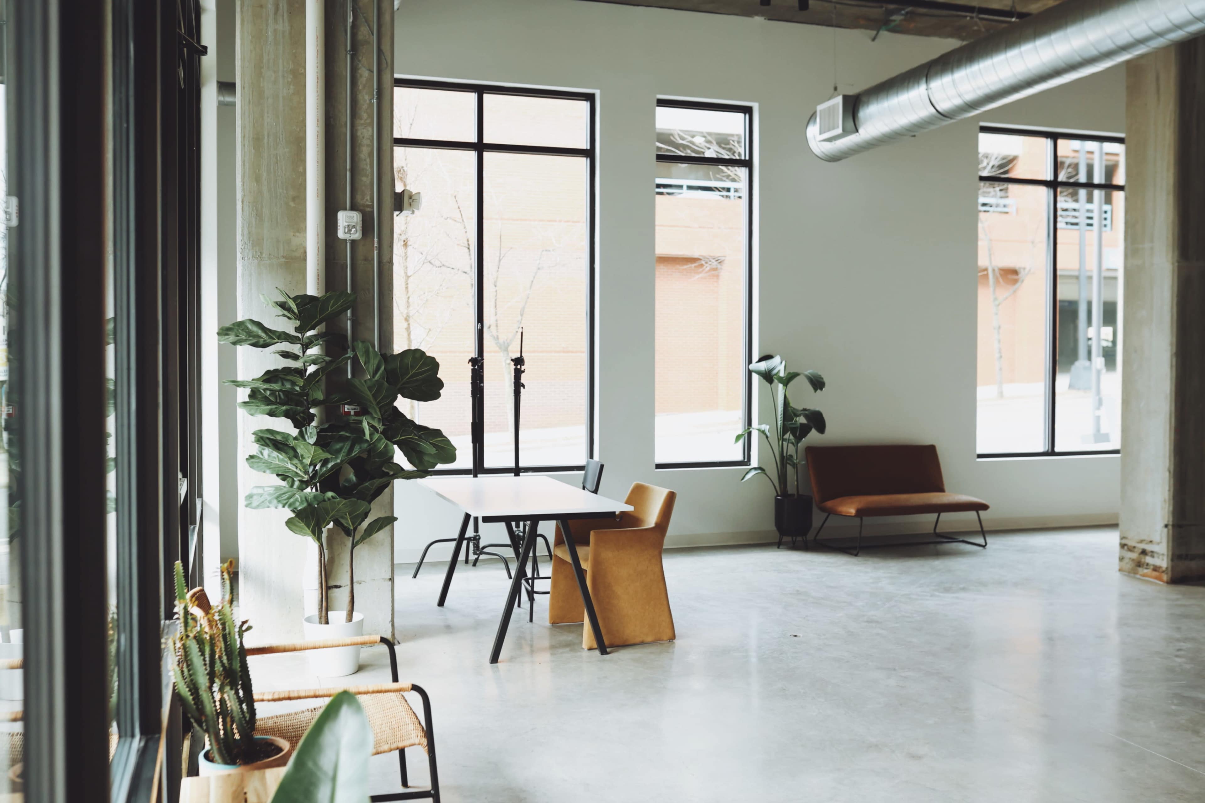 A modern interior space with large windows, a table and chairs, and plants, featuring concrete walls and flooring.